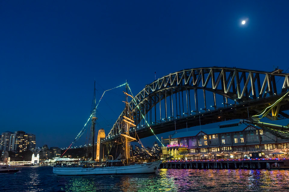 A sailboat illuminated by lights floats on water near a brightly lit bridge and a waterfront building at dusk.