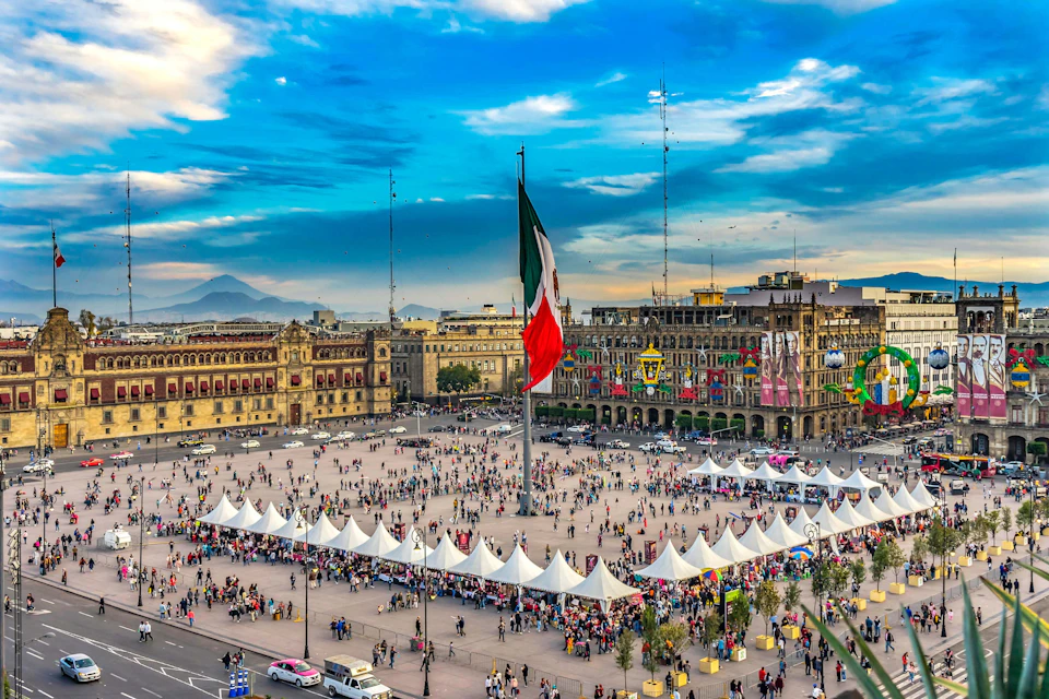Large plaza with white tents, a tall flagpole with the Mexican flag, surrounded by historic buildings and crowds of people.