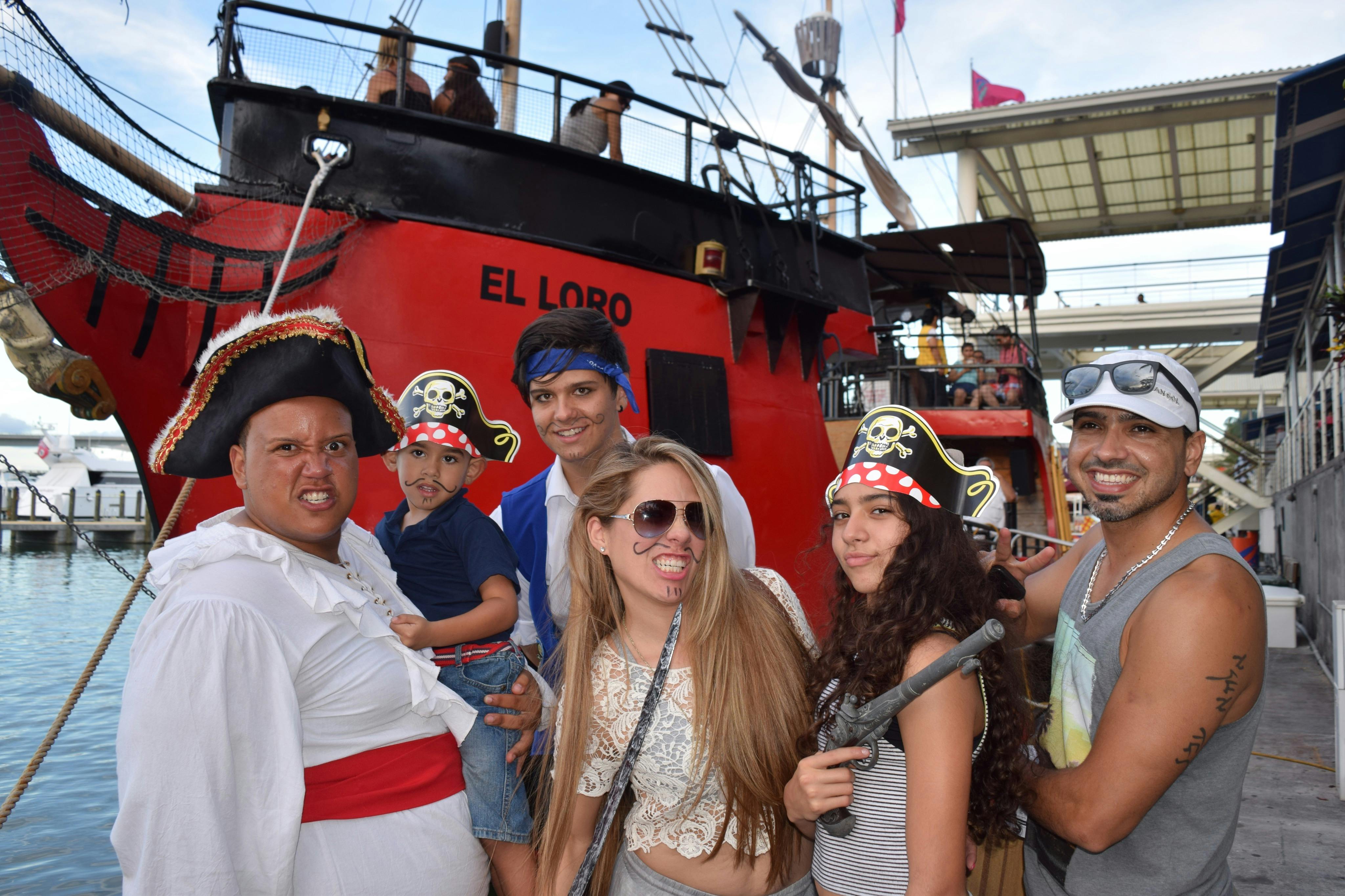 A group of people in pirate costumes smiling and posing in front of a red ship named 'El Loro' docked at a waterfront.
