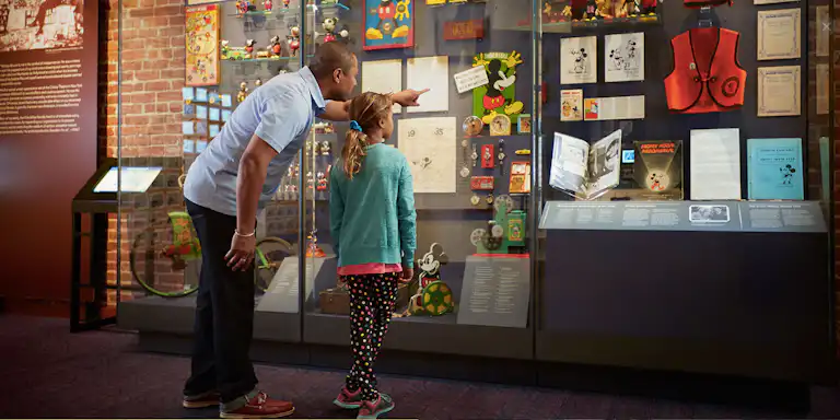 A man and a young girl examining a display case filled with various Mickey Mouse memorabilia in a museum.