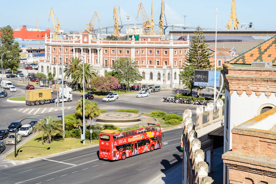 A red double-decker tour bus on a city street with palm trees, a large brick building, and industrial cranes in the background.