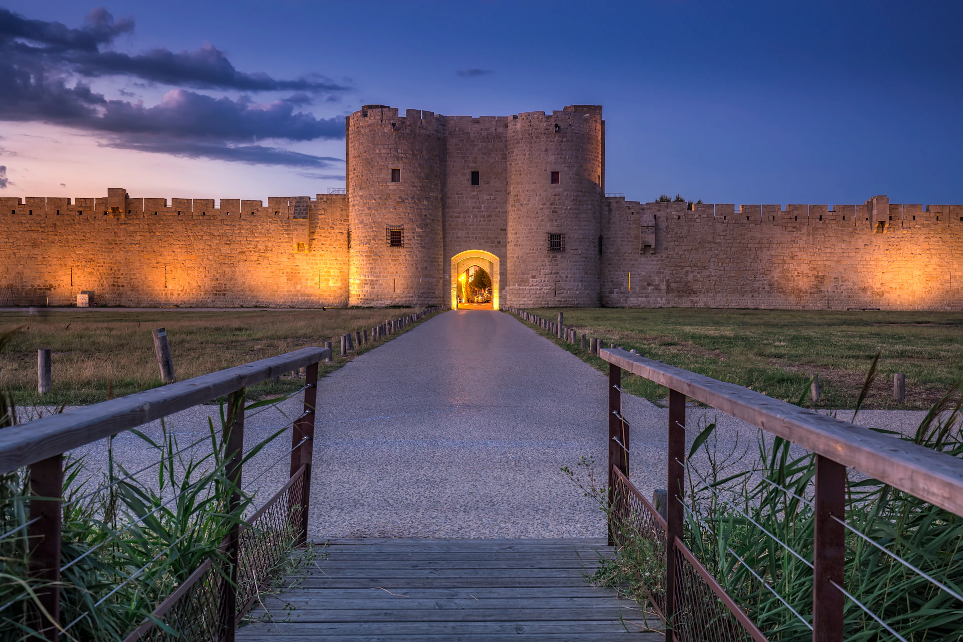 Stone castle with illuminated walls at dusk, viewed from a wooden bridge and path leading to the entrance.
