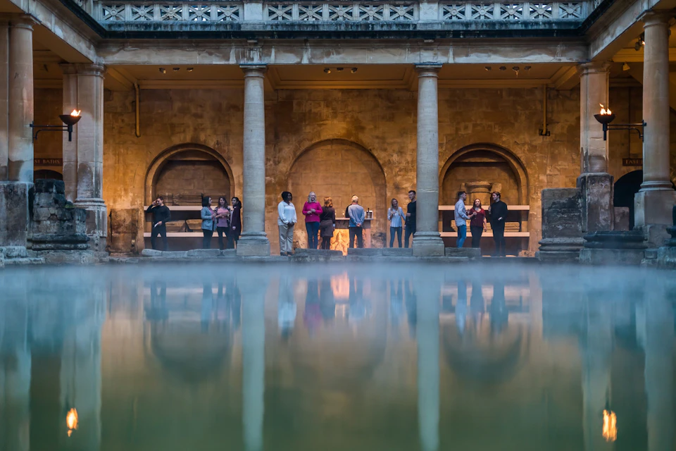 A group of people stand near stone arches and columns at the edge of a misty pool, with warm ambient lighting.