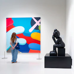 En person i vit skjorta och jeans tittar på en färgglad abstrakt målning på ett museum, bredvid en svart skulptur av en tänkande figur.