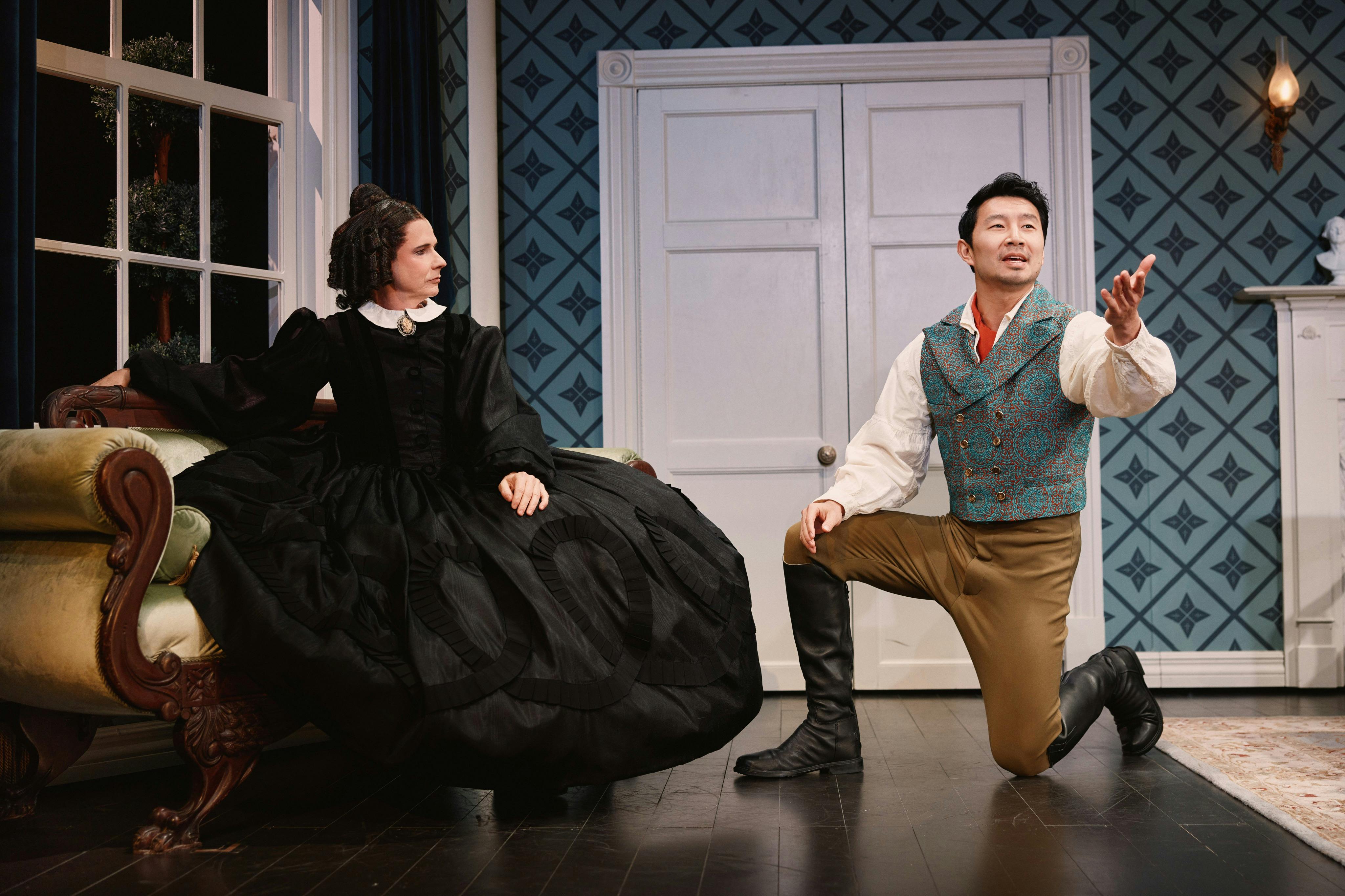 A man kneeling in a theatrical scene, dressed in period costume, with a patterned vest and boots, gesturing with his hand.