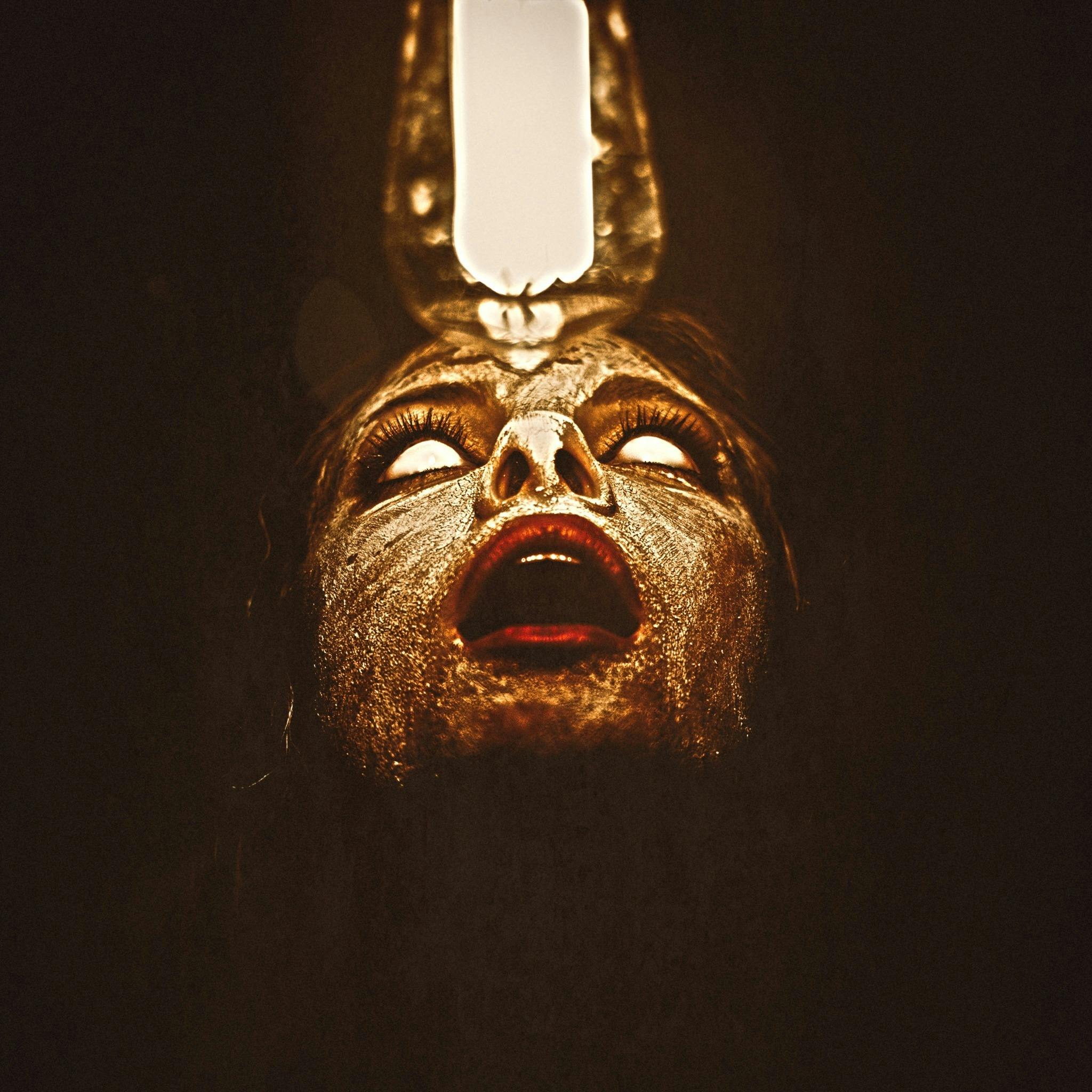 A person with white eyes and an open mouth, illuminated from above, their face adorned with tribal-like paint against a dark background.