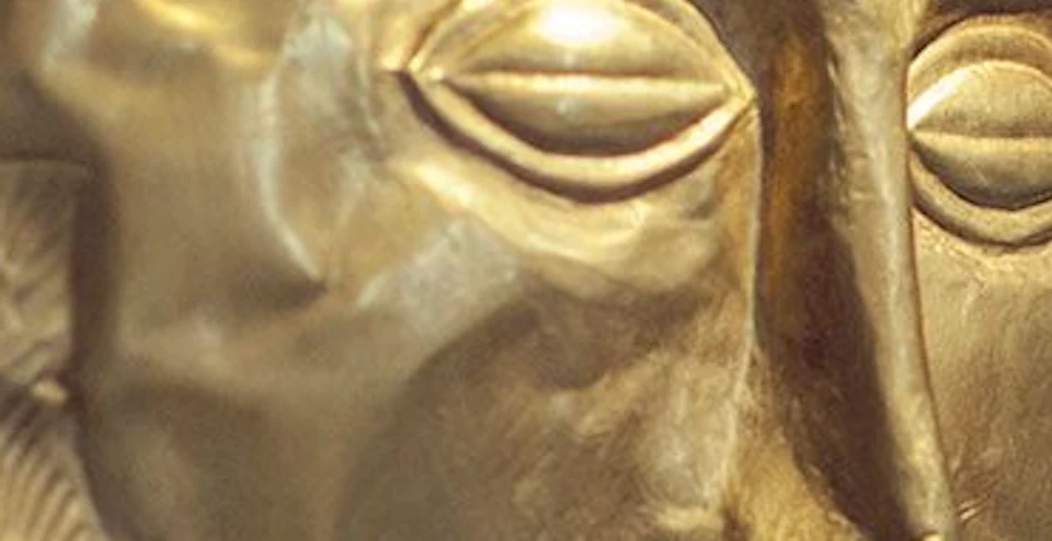An ancient gold funerary mask with detailed facial features, displayed on a dark wall.