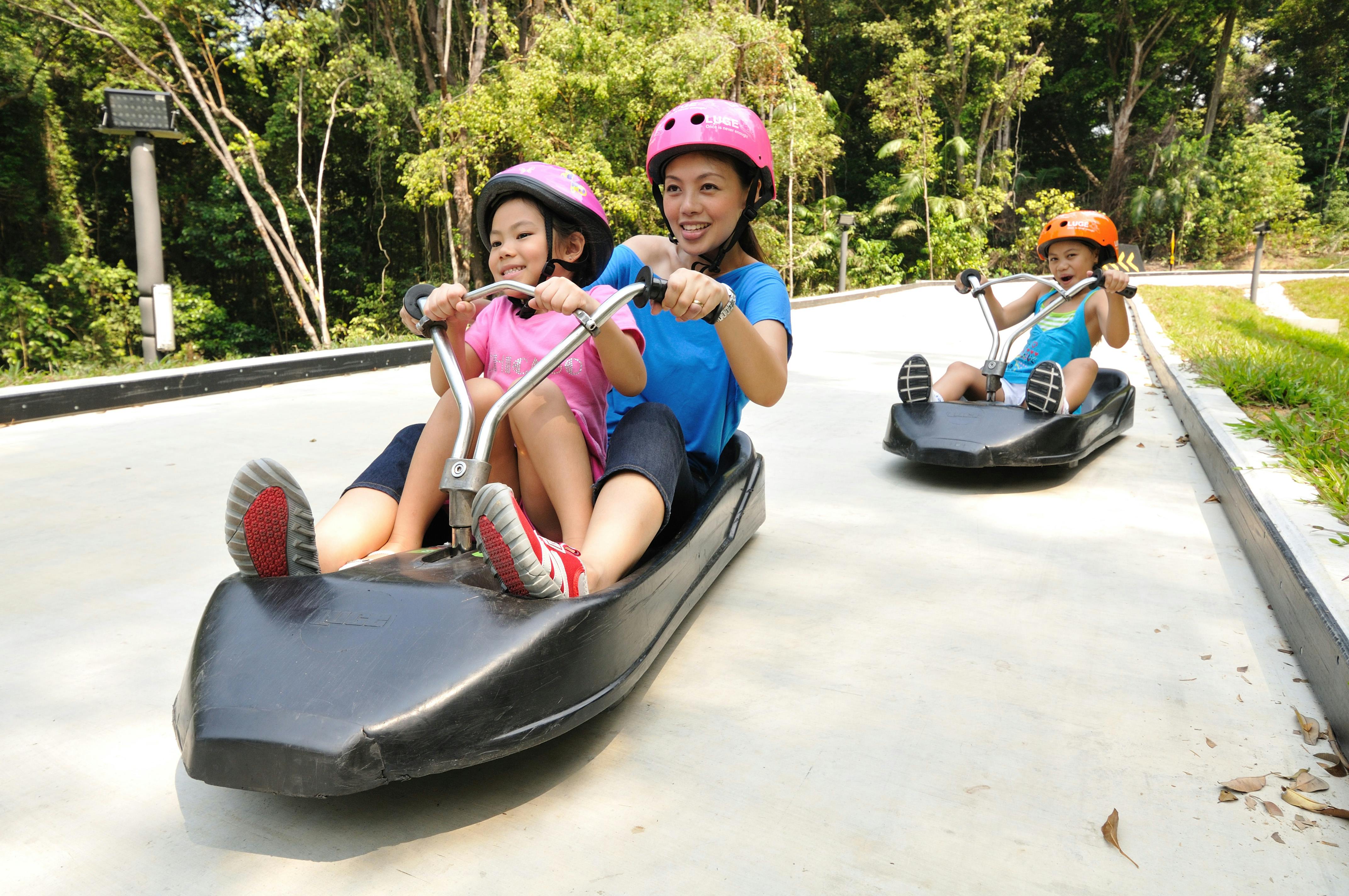 Three people on luges, two wearing pink helmets and one wearing an orange helmet, riding on a track surrounded by trees.