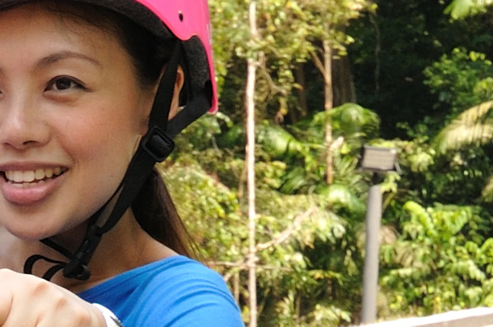 Three people on luges, two wearing pink helmets and one wearing an orange helmet, riding on a track surrounded by trees.