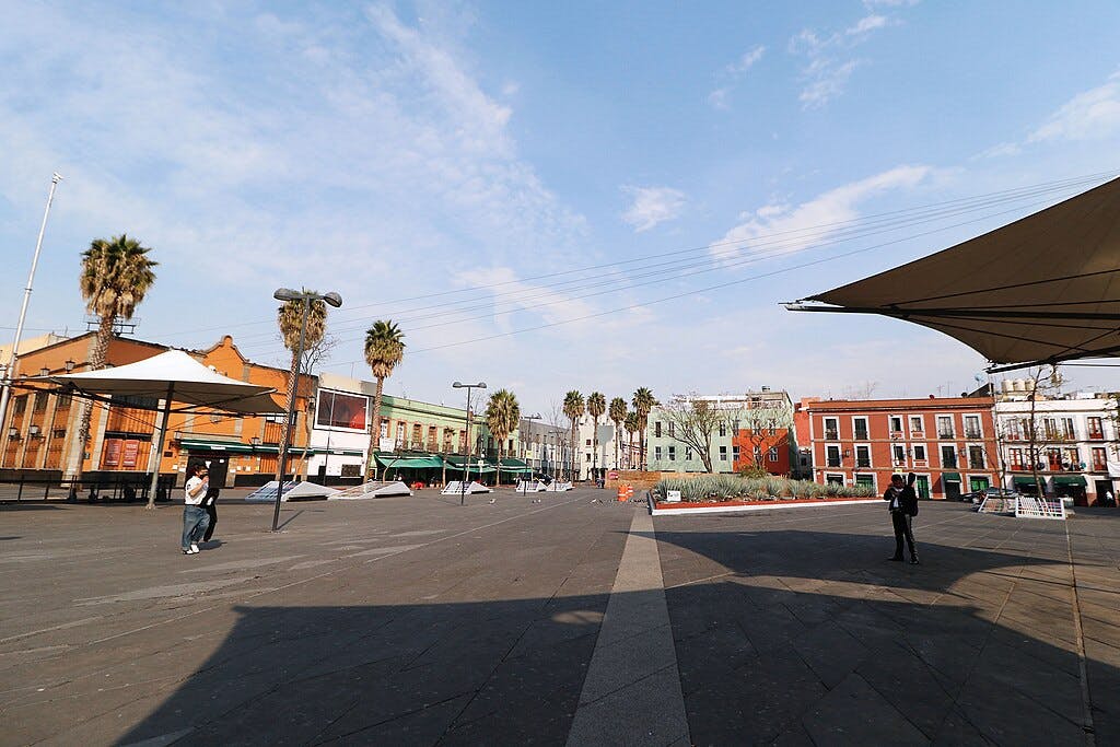 Open plaza with scattered trees and people, surrounded by colorful buildings under a blue sky with light clouds.