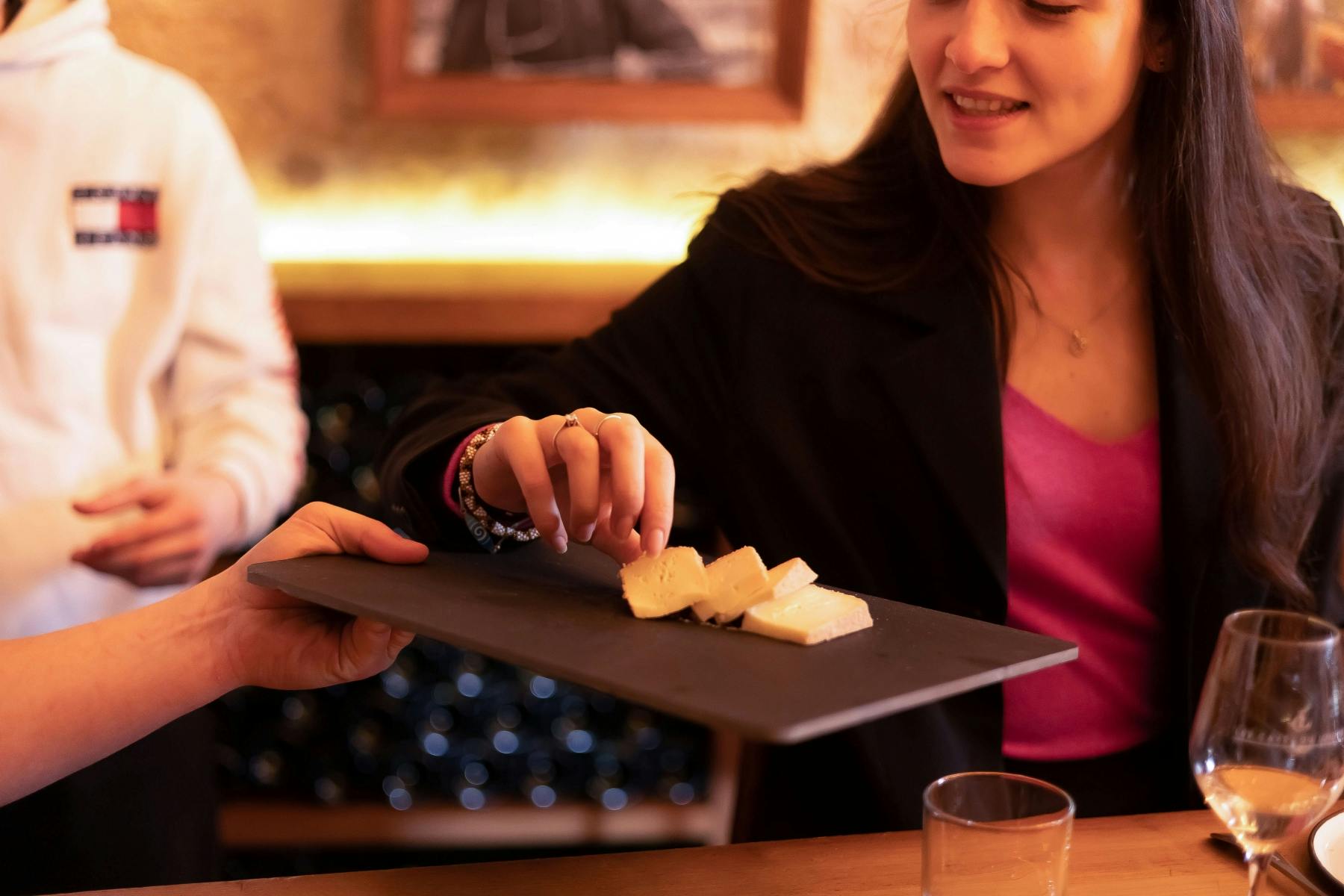 Женщина в черном пиджаке держит шифер с ломтиками сыра. На деревянном столе виден бокал для вина и тарелка.
