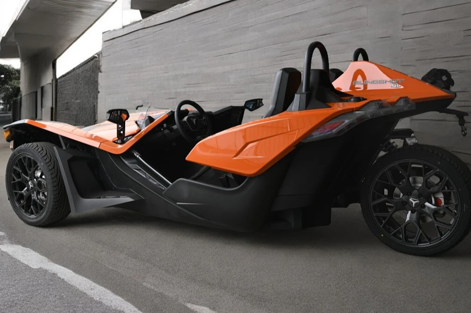 A bright orange, futuristic-looking three-wheeled vehicle parked on a paved surface near a concrete wall.
