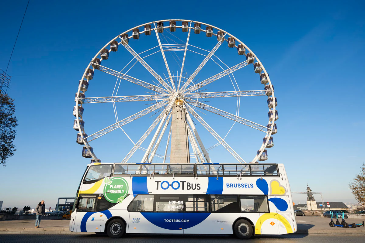 A double-decker tour bus labeled "TOOTBus Brussels" parked in front of a large Ferris wheel against a clear blue sky.