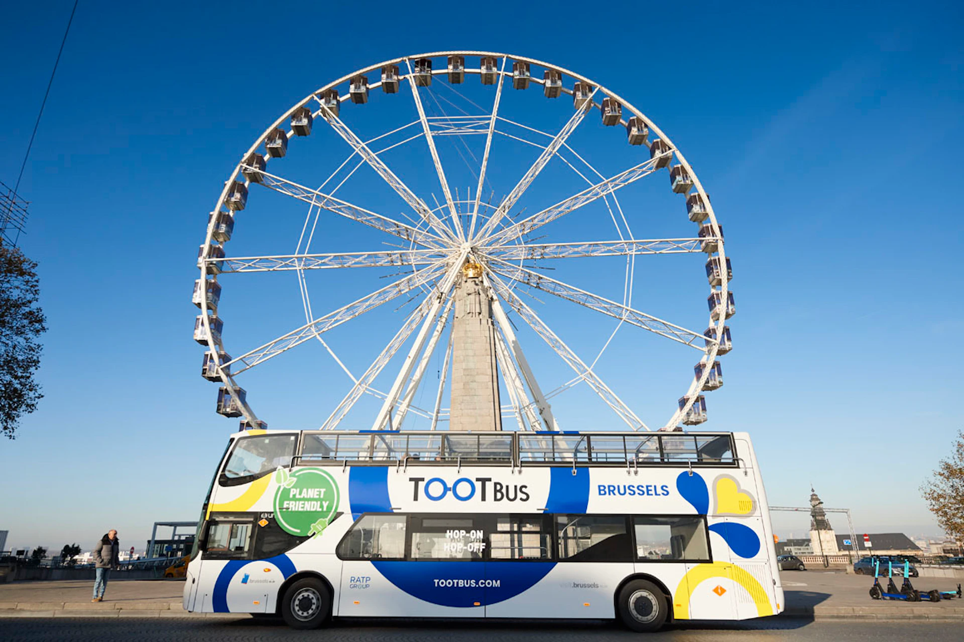 A double-decker tour bus labeled "TOOTBus Brussels" parked in front of a large Ferris wheel against a clear blue sky.