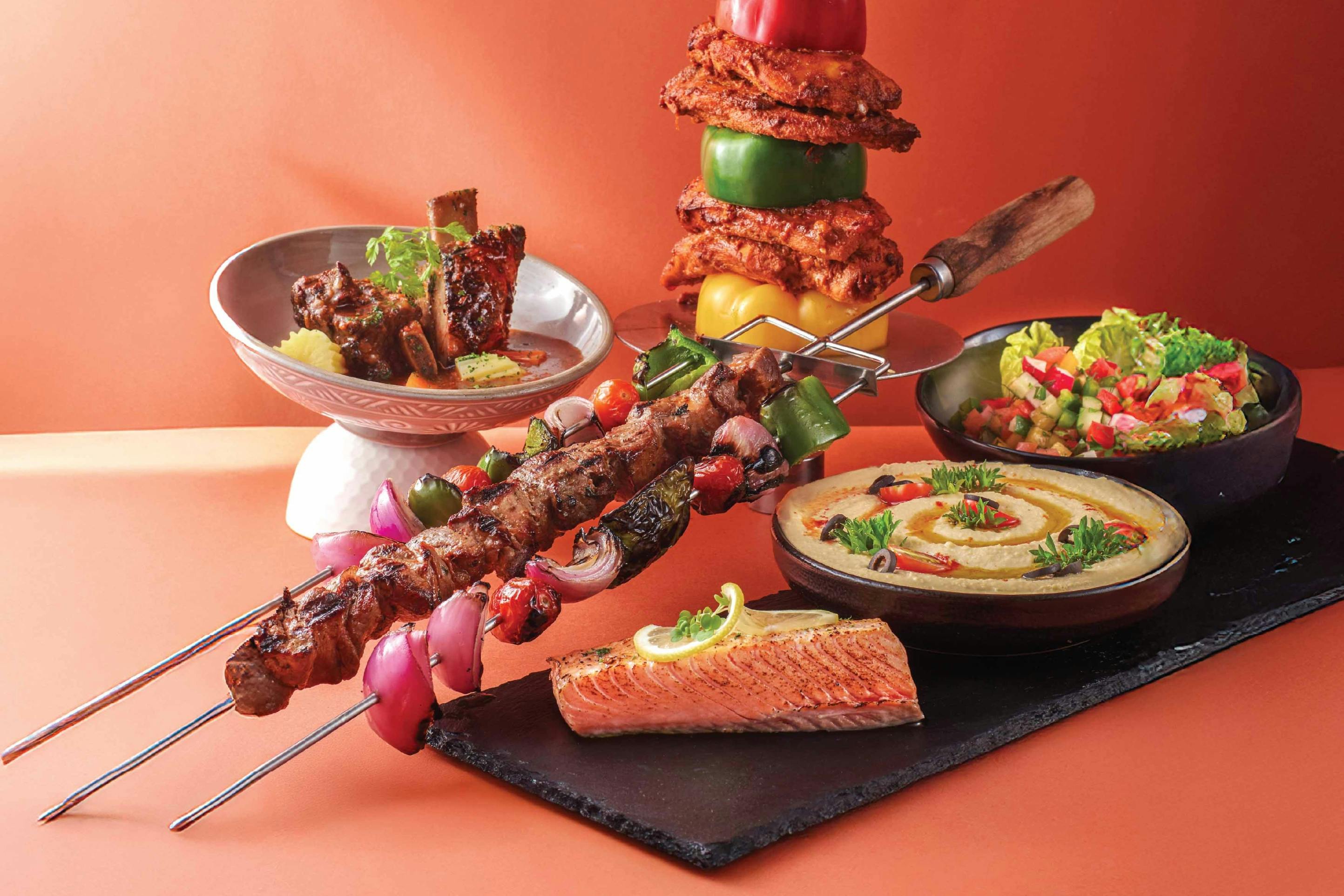 An assortment of dishes including skewered meat, grilled salmon, hummus, guacamole, ribs, and a vegetable stack.