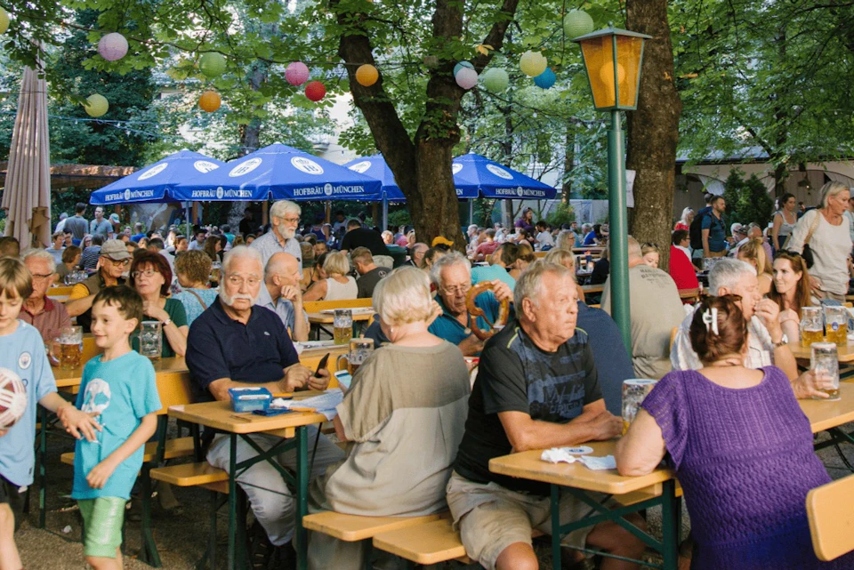 A lively outdoor beer garden with people sitting at tables under trees and umbrellas, enjoying food and drinks.
