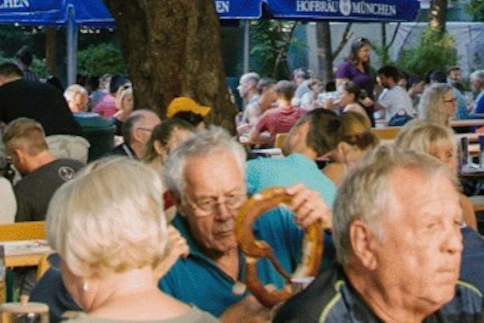 A lively outdoor beer garden with people sitting at tables under trees and umbrellas, enjoying food and drinks.
