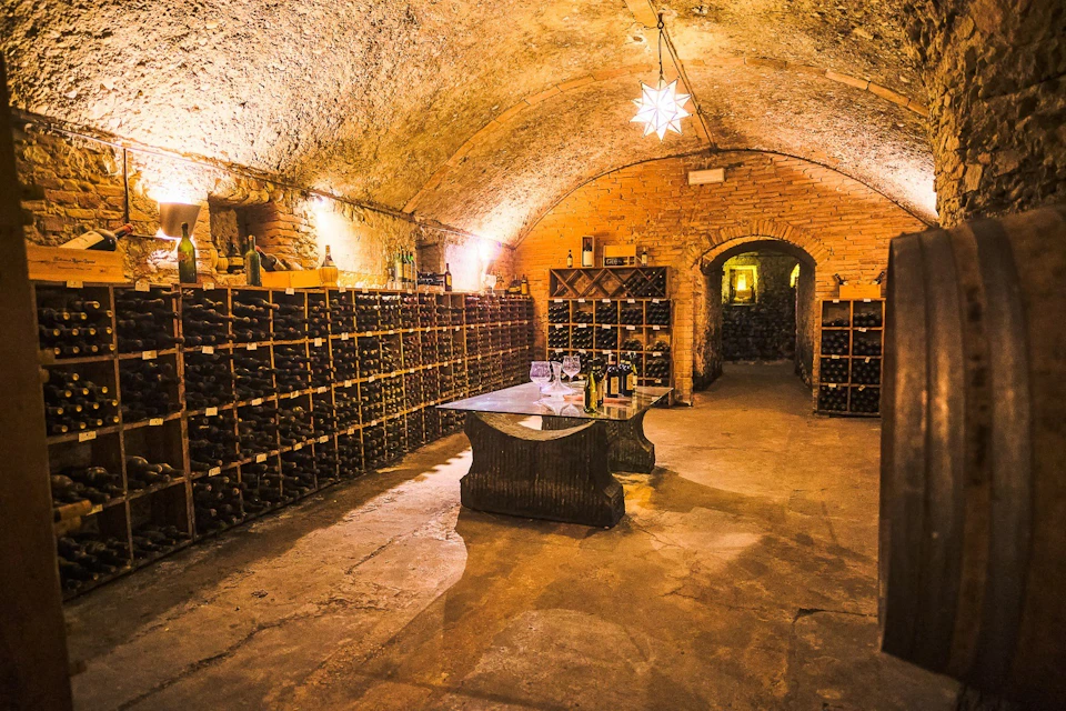 A warmly lit wine cellar with arched stone ceilings, wooden wine racks, a table, and bottles on display.