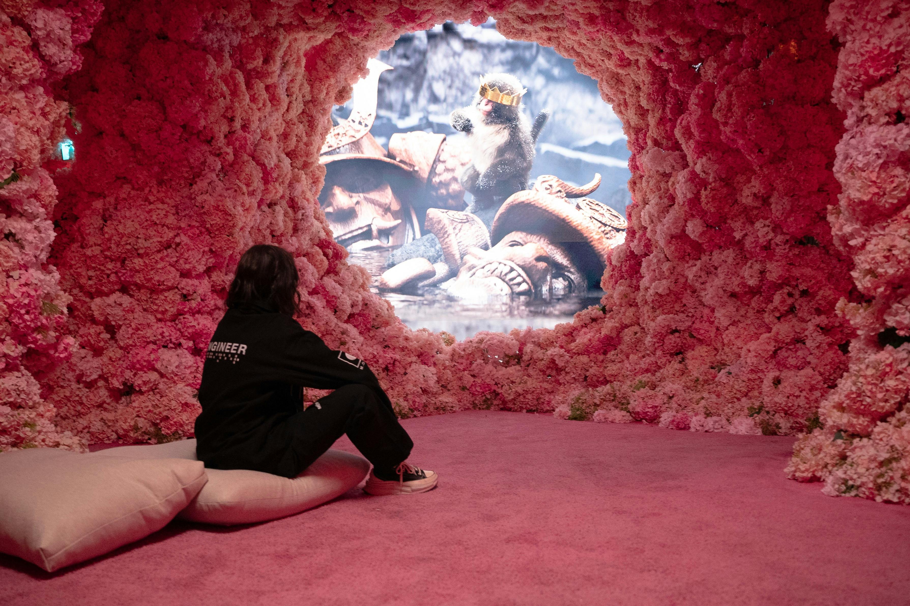 분홍색 쿠션에 앉아 기발한 장면을 보여주는 꽃무늬 아치길을 통해 화면을 보고 있는 어두운 옷을 입은 사람.