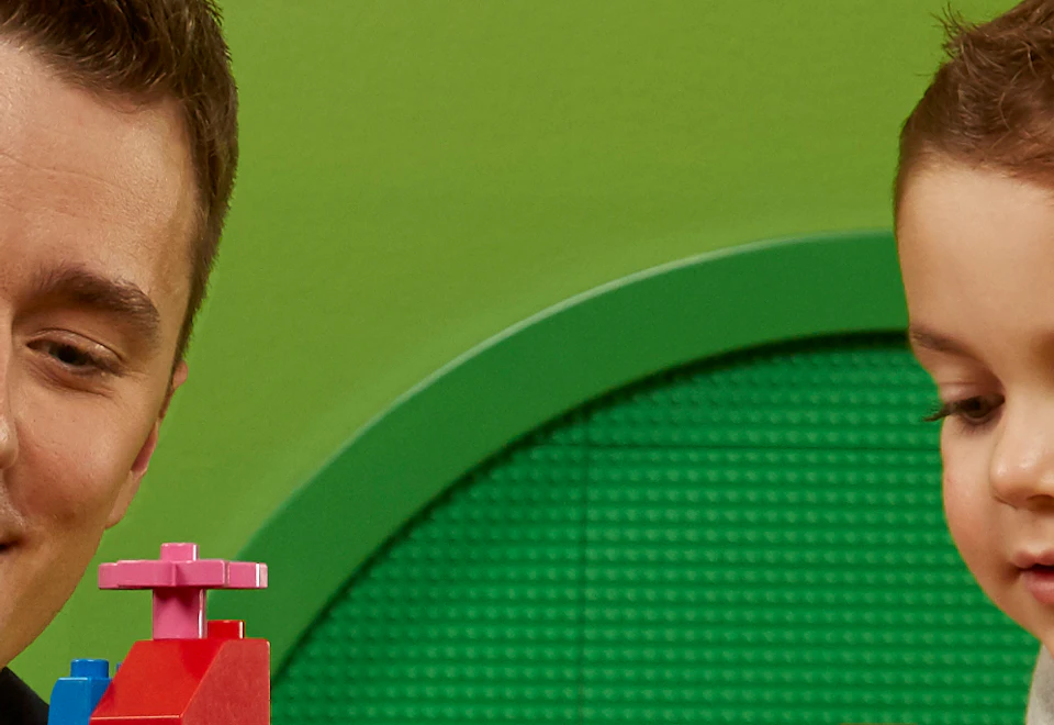 A man and a boy are building a colorful structure with LEGO bricks on a yellow table against a green background with LEGO pieces.