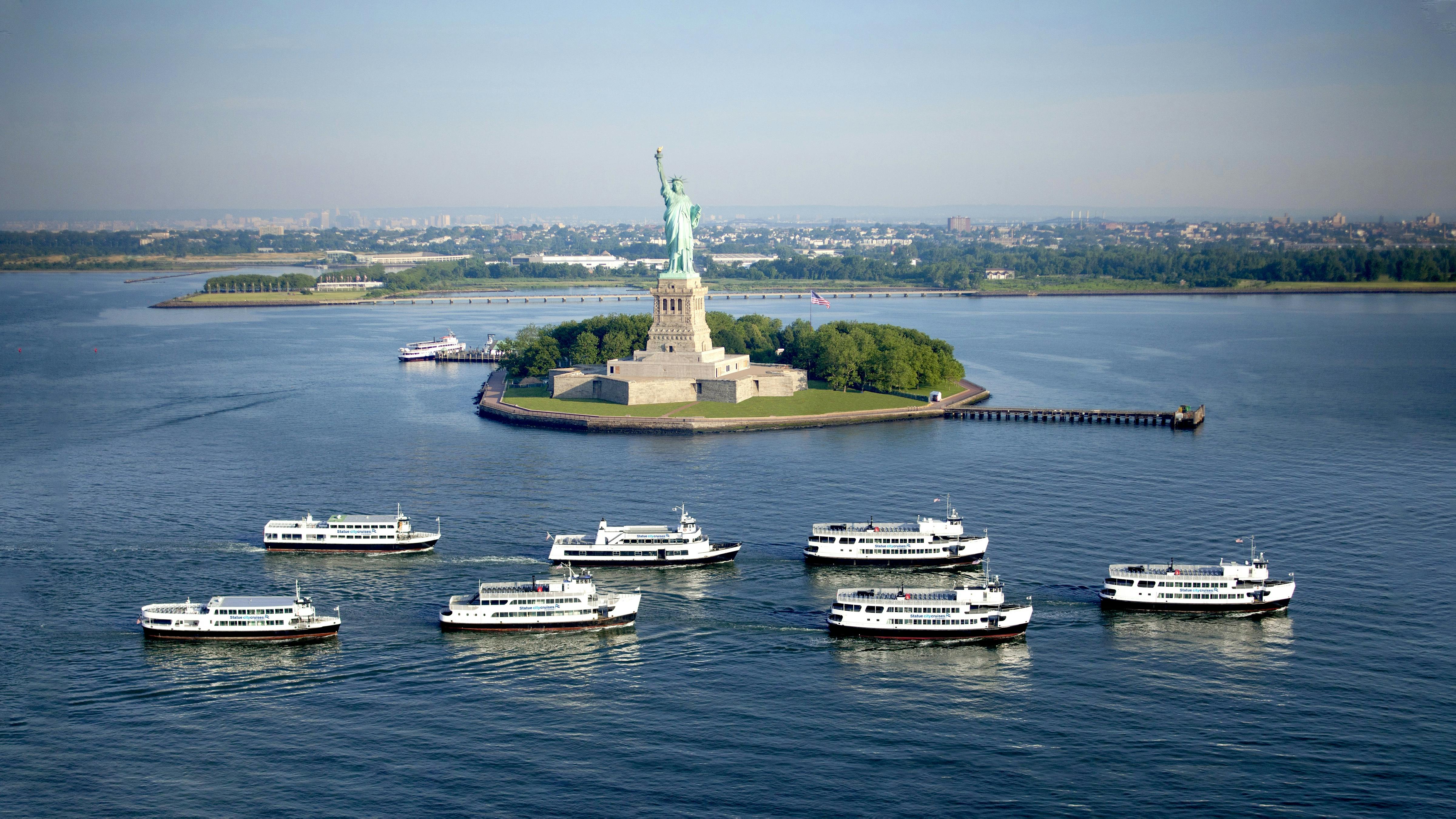 Frihedsgudinden på en ø med træer, omgivet af vand, og flere færger i forgrunden.
