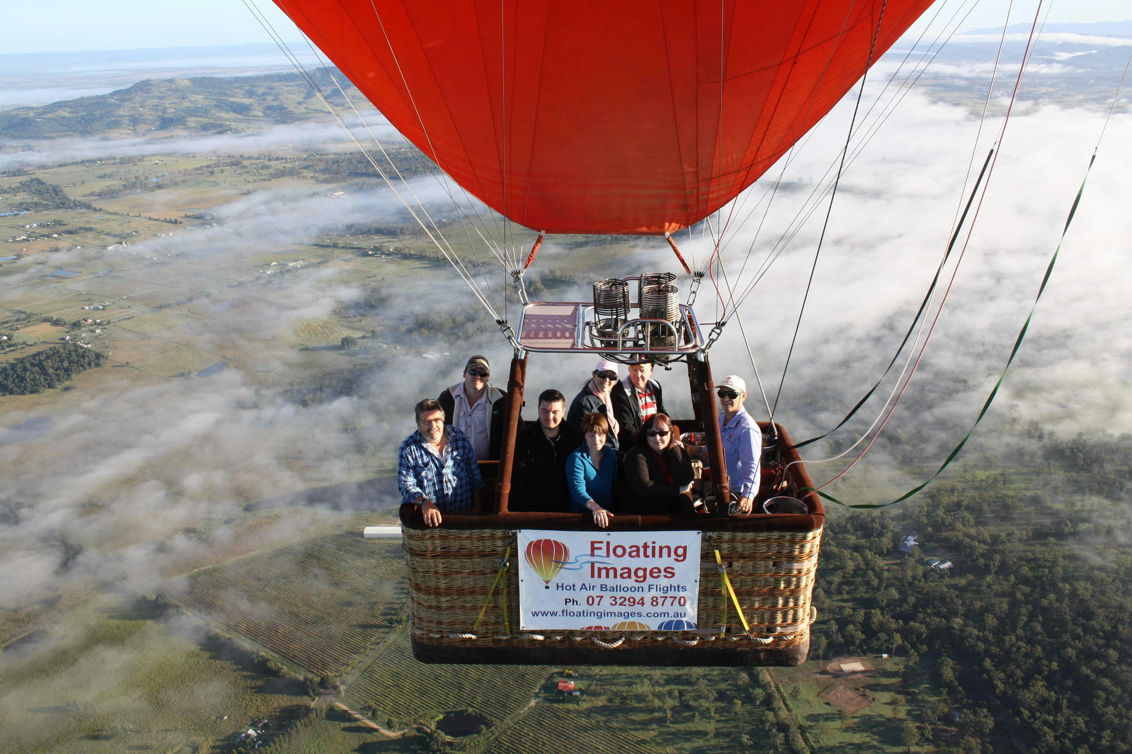 Un grup de persones en un globus aerostàtic amb una marquesina vermella, flotant sobre un paisatge pintoresc i boirós.
