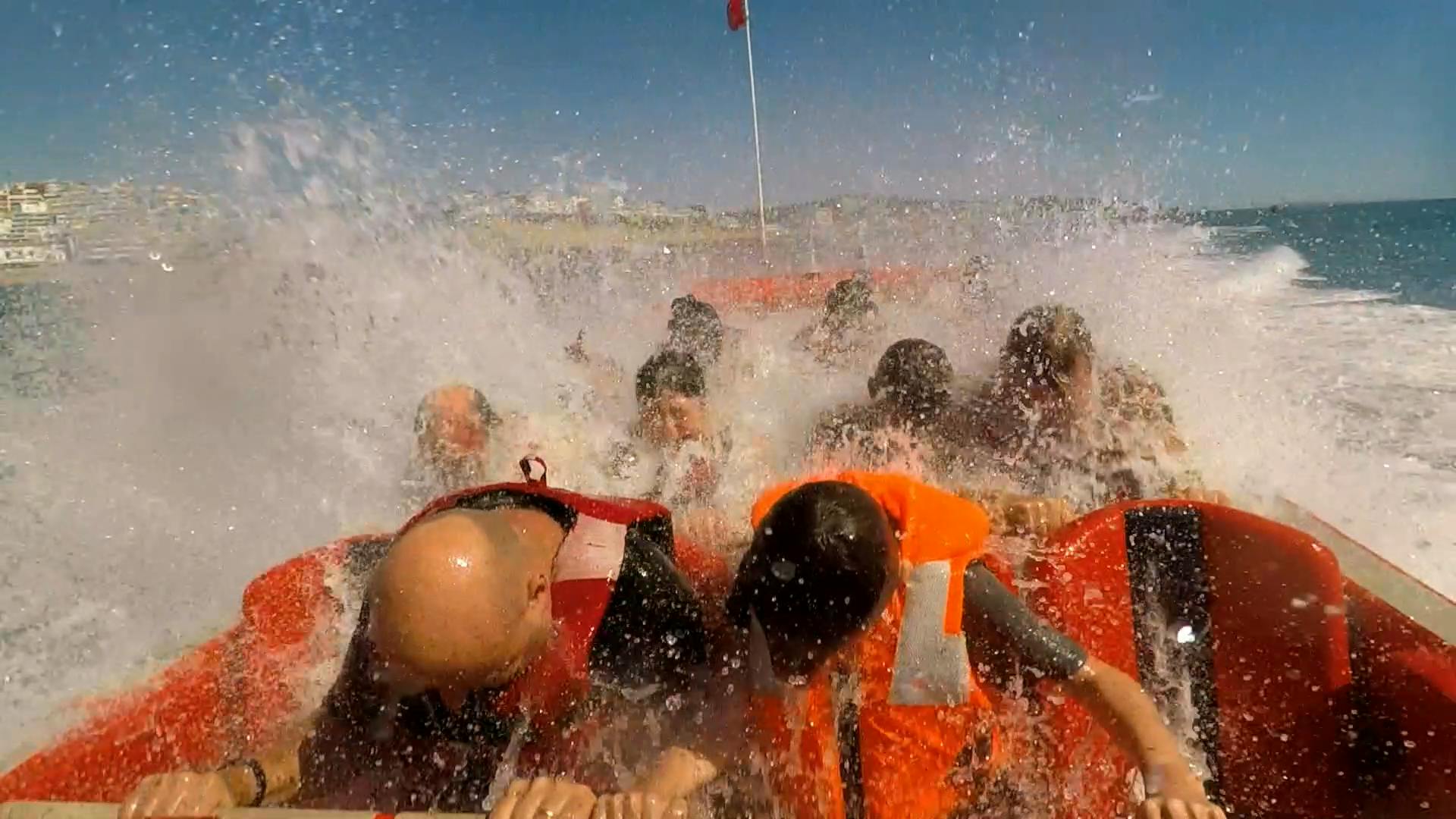 Skupina lidí v oranžových záchranných vestách na lodi, která je postříkána velkými vlnami pod jasně modrým nebem.