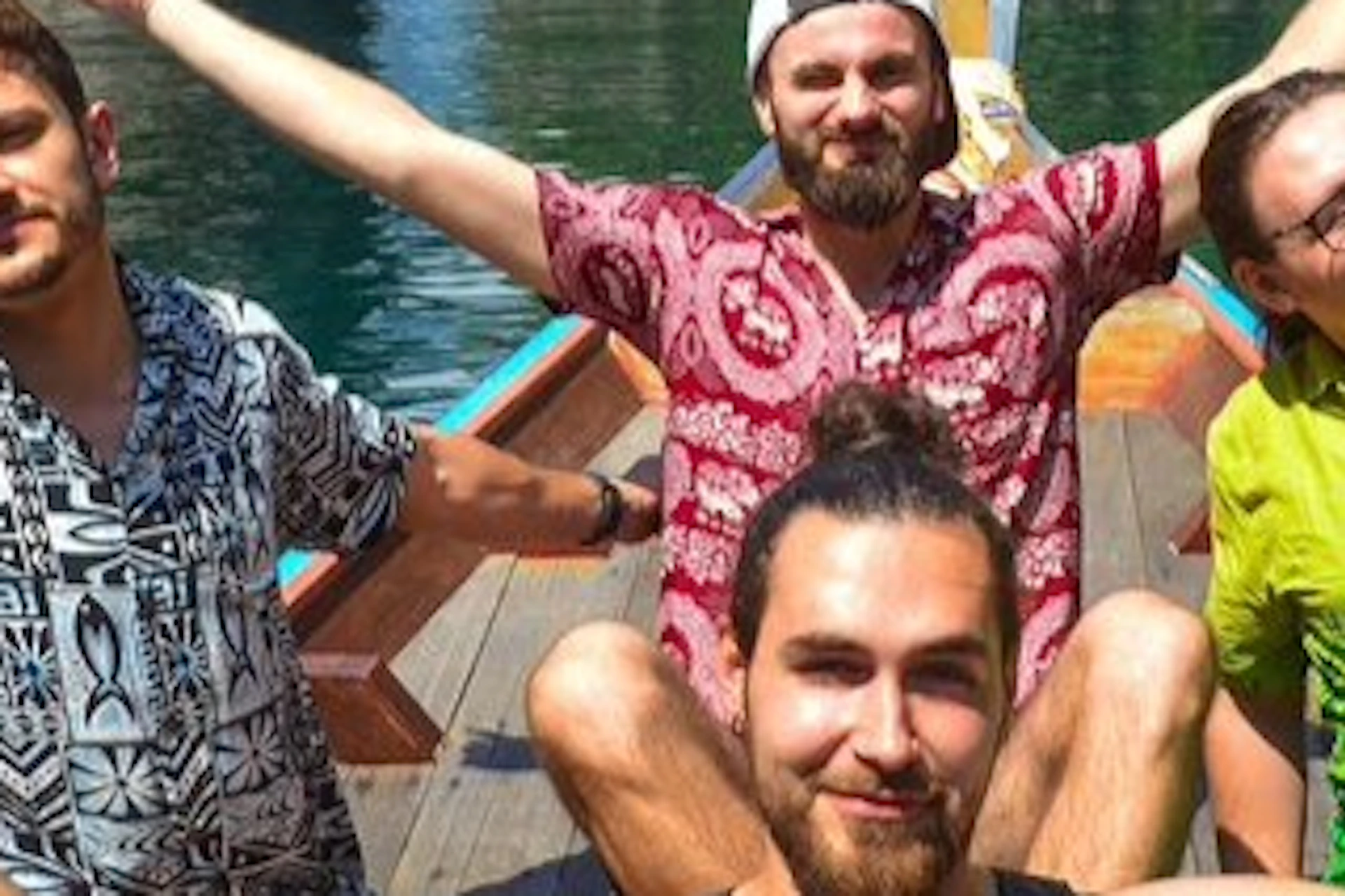 Four men with outstretched arms sit in a boat on a turquoise lake, surrounded by tall, lush green islands under a blue sky.