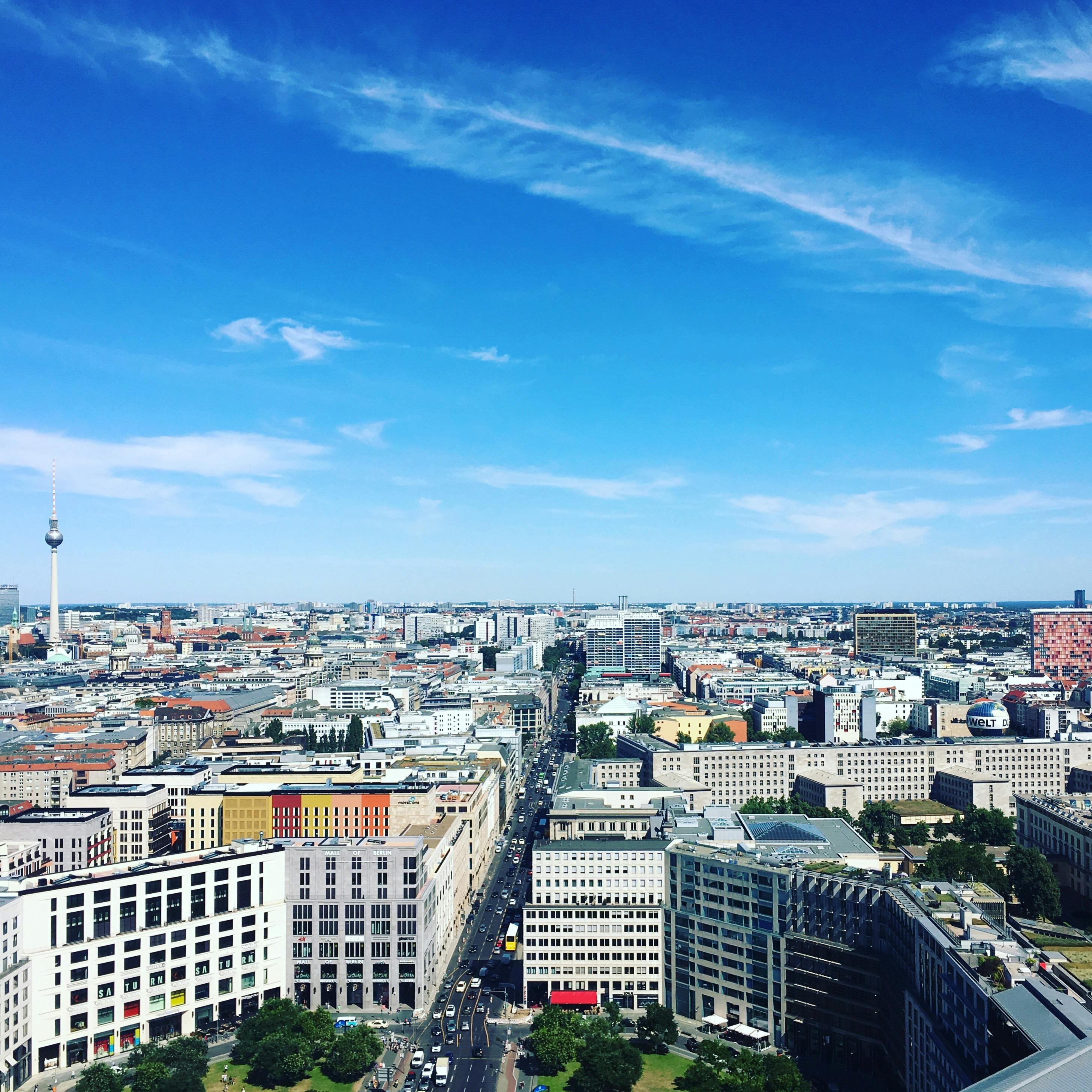 Luchtfoto van een stadsgezicht met moderne gebouwen, groene ruimtes, een hoge toren en een drukke straat onder een blauwe lucht met verspreide wolken.