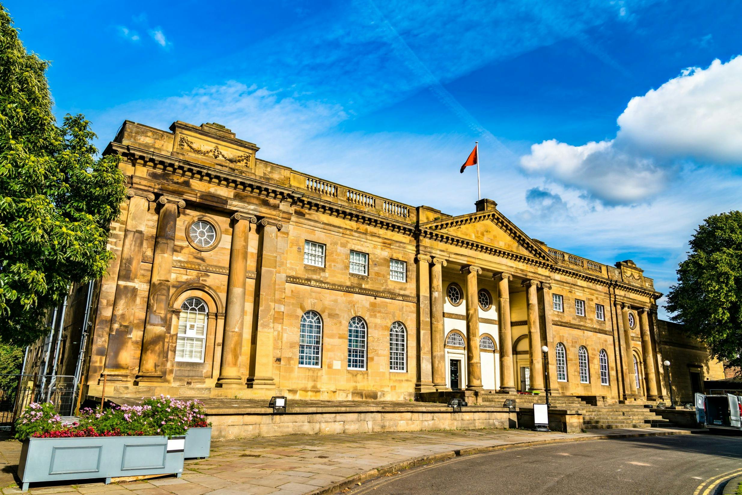 Zabytkowy kamienny budynek z kolumnami, łukowatymi oknami i czerwoną flagą na górze, pod błękitnym niebem z chmurami.