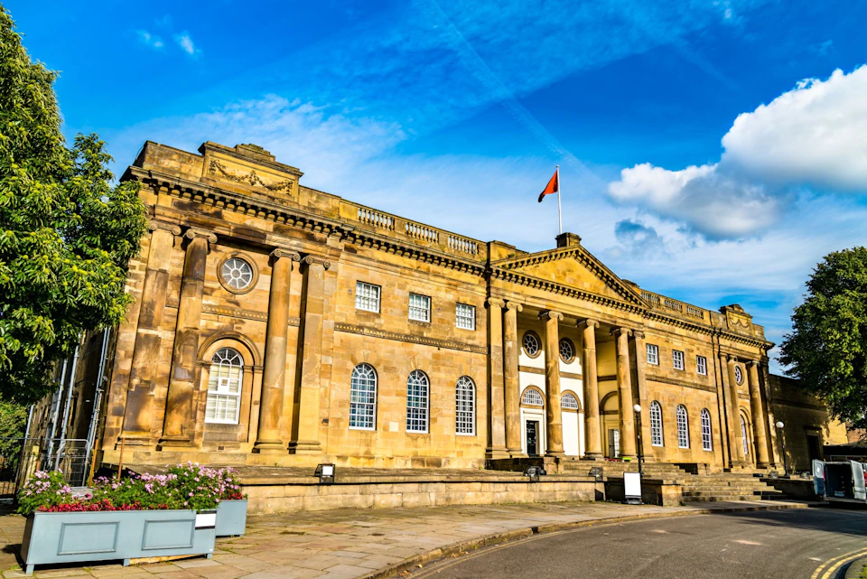 Historic stone building with columns, arched windows, and a red flag on top, under a blue sky with clouds.