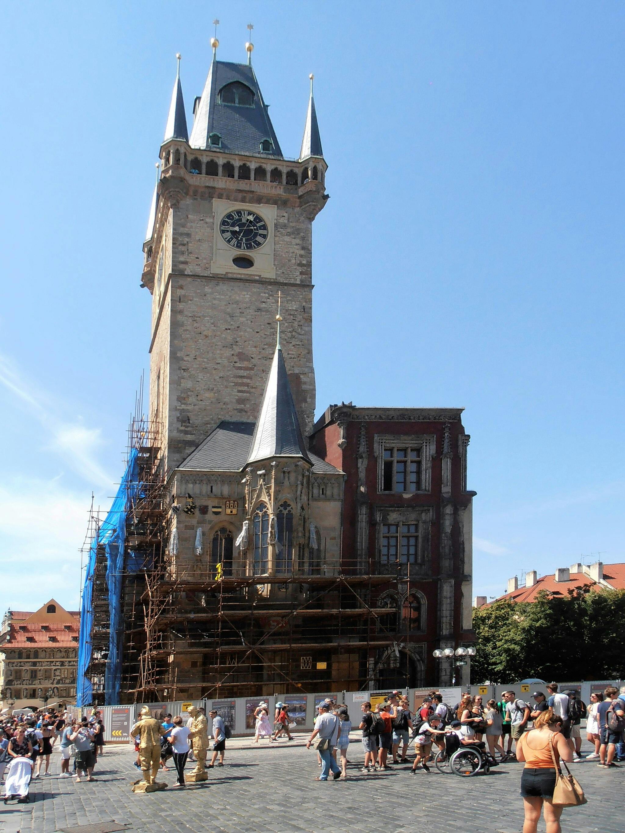 People gather near a tall historic clock tower with scaffolding, under a clear blue sky.