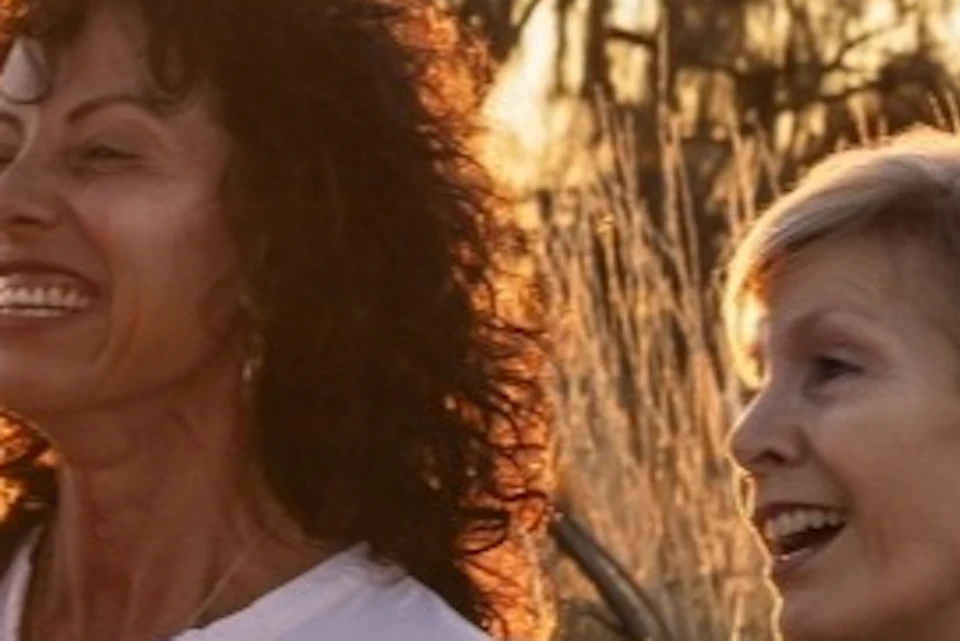 Three people stand and laugh in a field at sunset, with tall grass and trees in the background.