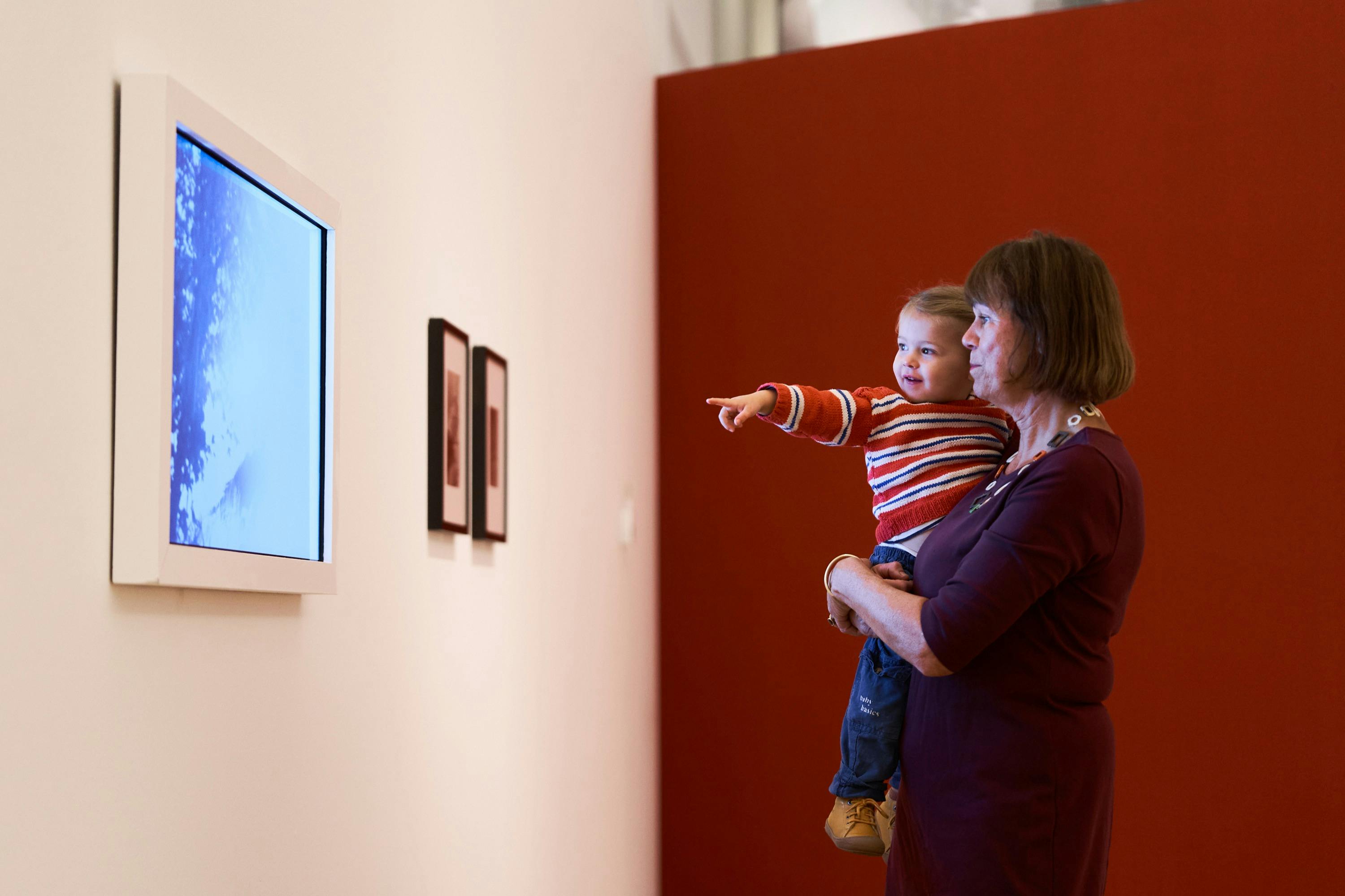 Μια ηλικιωμένη γυναίκα κρατά ένα μικρό παιδί που δείχνει μια κορνιζαρισμένη τέχνη σε έναν τοίχο με κόκκινο φόντο.