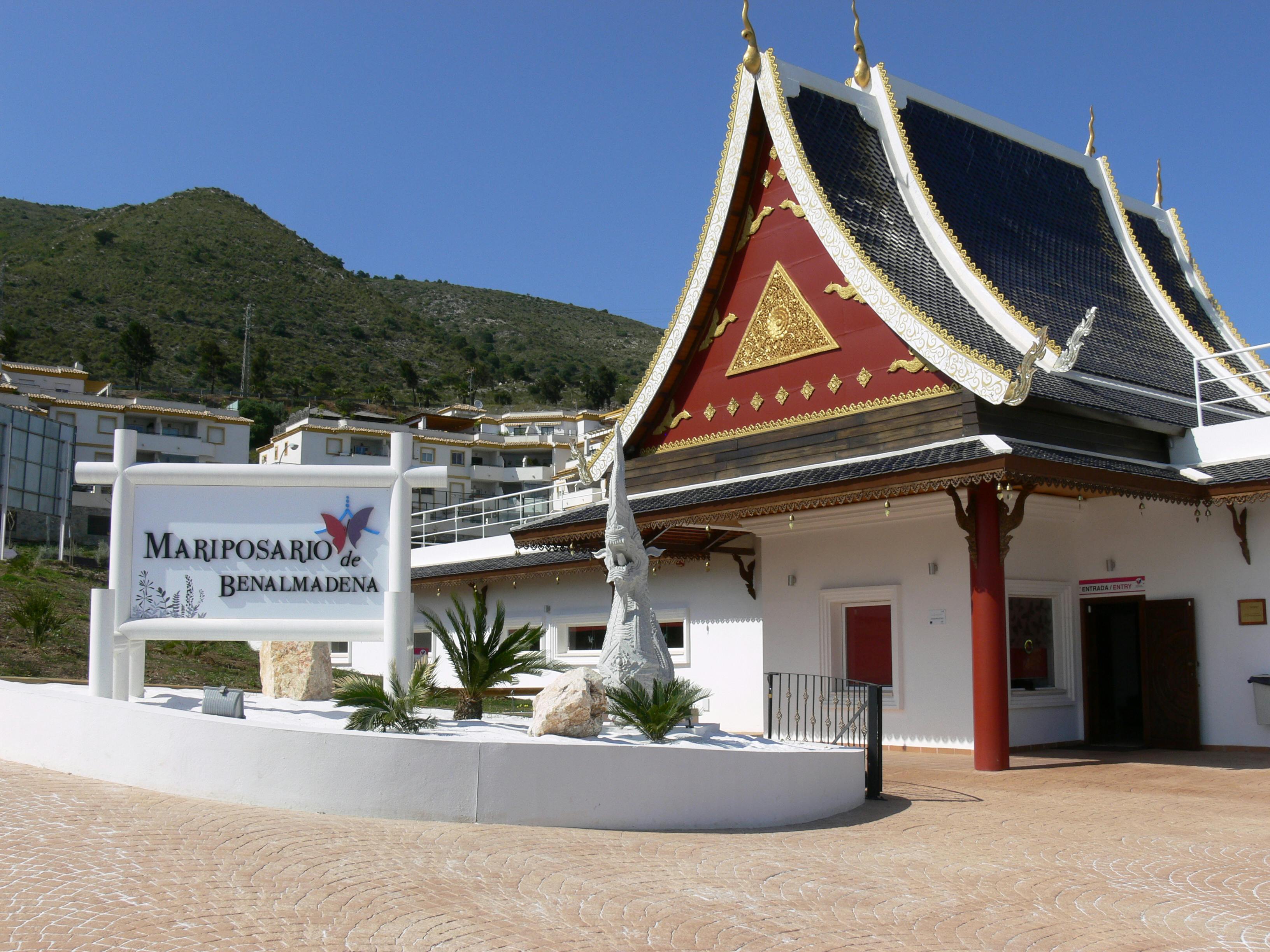 Byggnad i thailändsk stil med utsmyckat tak, skylt med texten &#34;Mariposario de Benalmadena&#34;, växter och bergig bakgrund under en klarblå himmel.