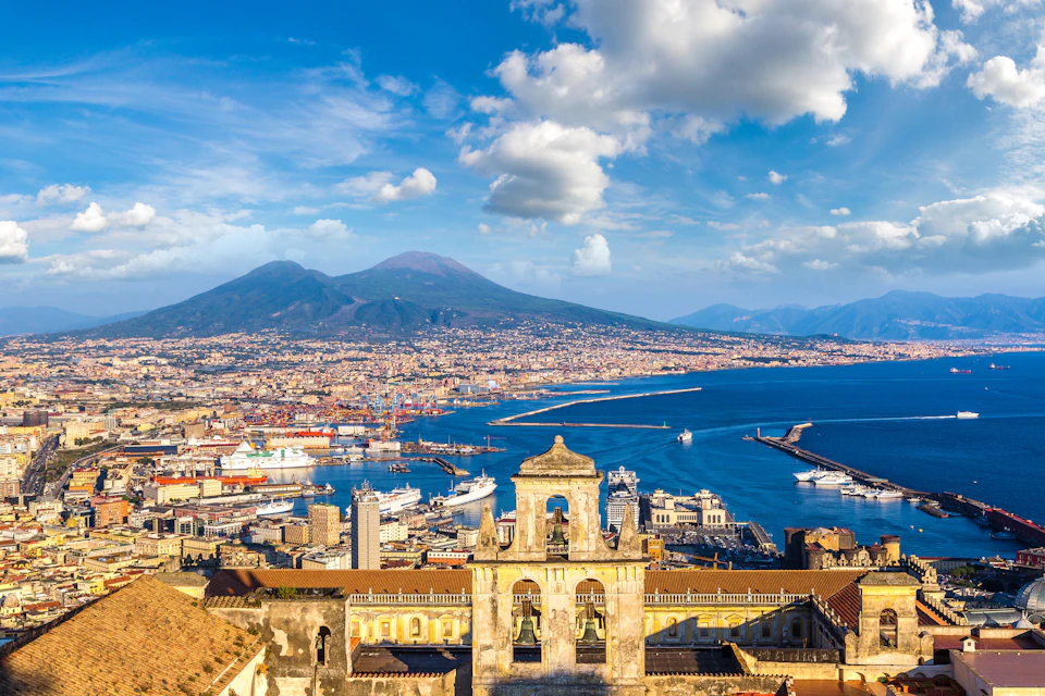 Panoramic view of a coastal city with a large mountain in the background, a harbor with ships, and a historic bell tower in the foreground.
