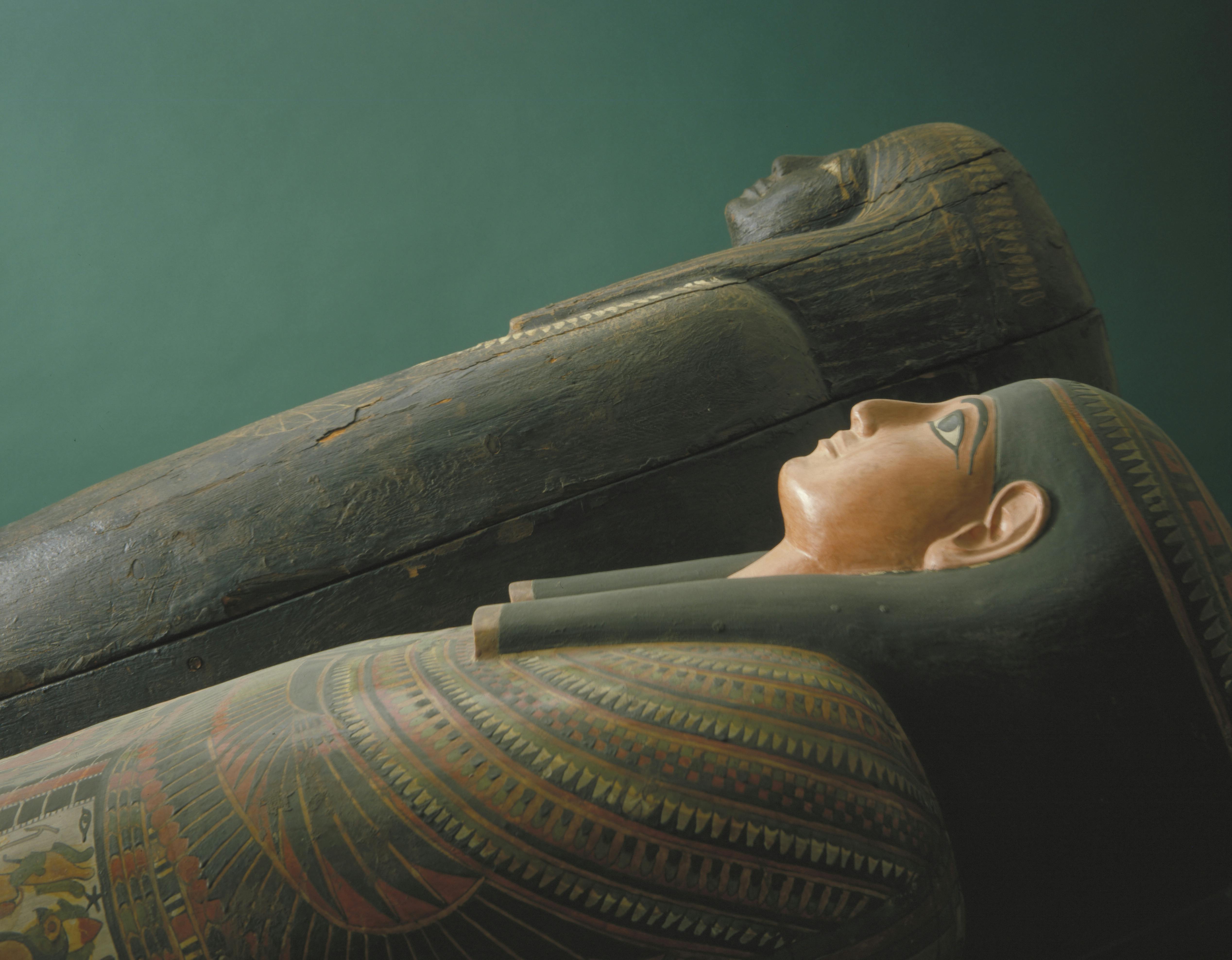 Due antichi sarcofagi egizi, uno dipinto con motivi dettagliati e un volto umano, l'altro scuro e semplice, giacciono uno accanto all'altro.