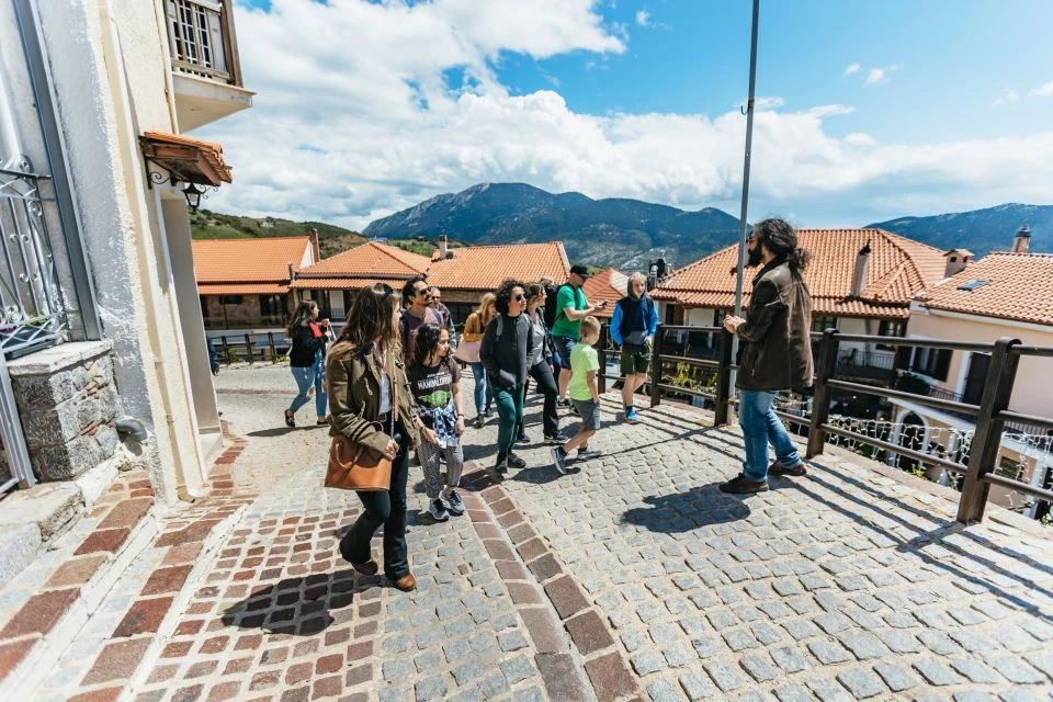 Tourists walking on a cobblestone street with a backdrop of red-roofed buildings and mountains under a blue sky.