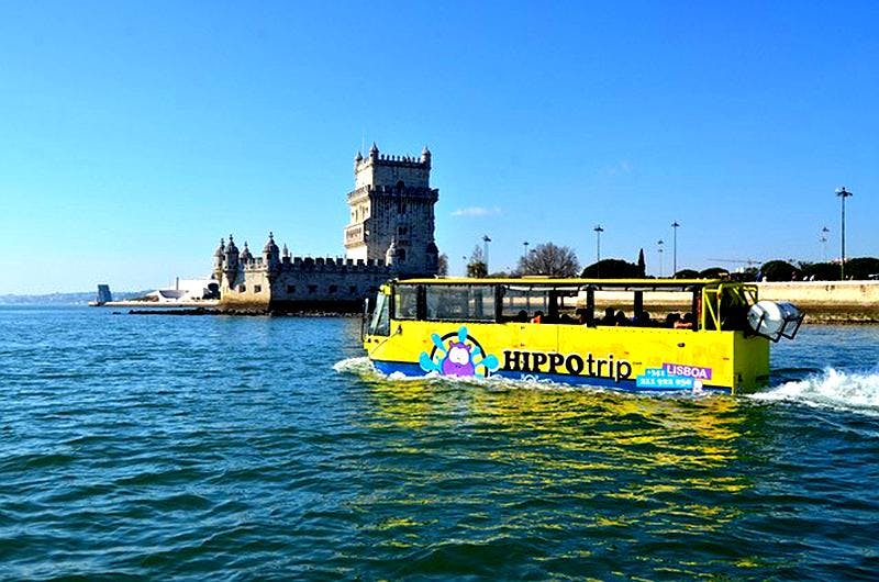 A yellow amphibious tour bus sails near an old stone tower on a clear day with a blue sky. The bus has "HIPPOtrip" branding.