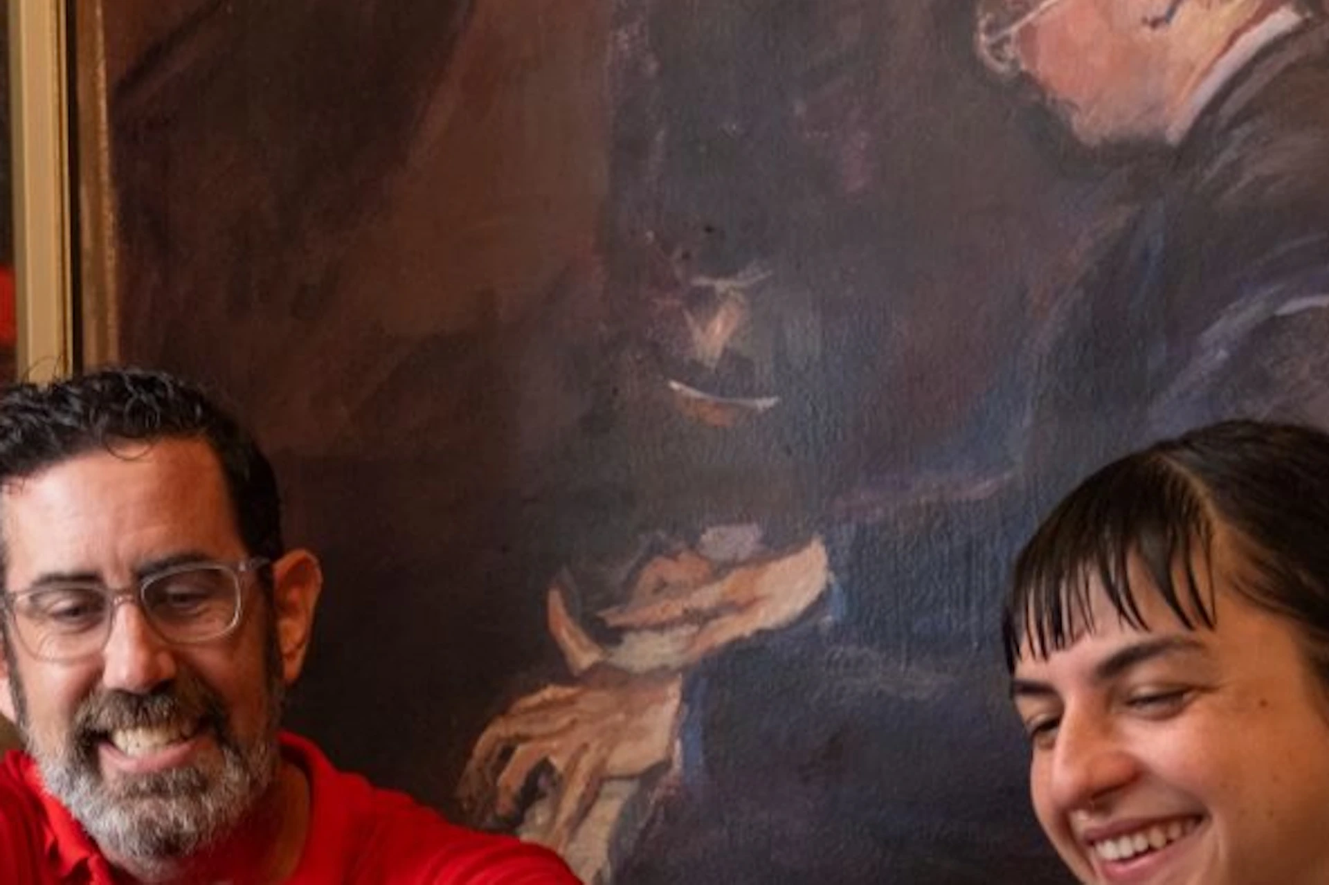 A man and woman enjoy a meal at a rustic restaurant, sharing sandwiches and fries, with coffee cups and a painting in the background.
