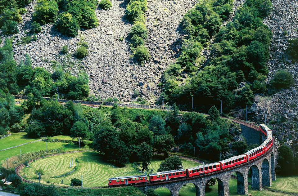 A red train travels on a stone viaduct through a lush, green valley surrounded by rocky hillsides.
