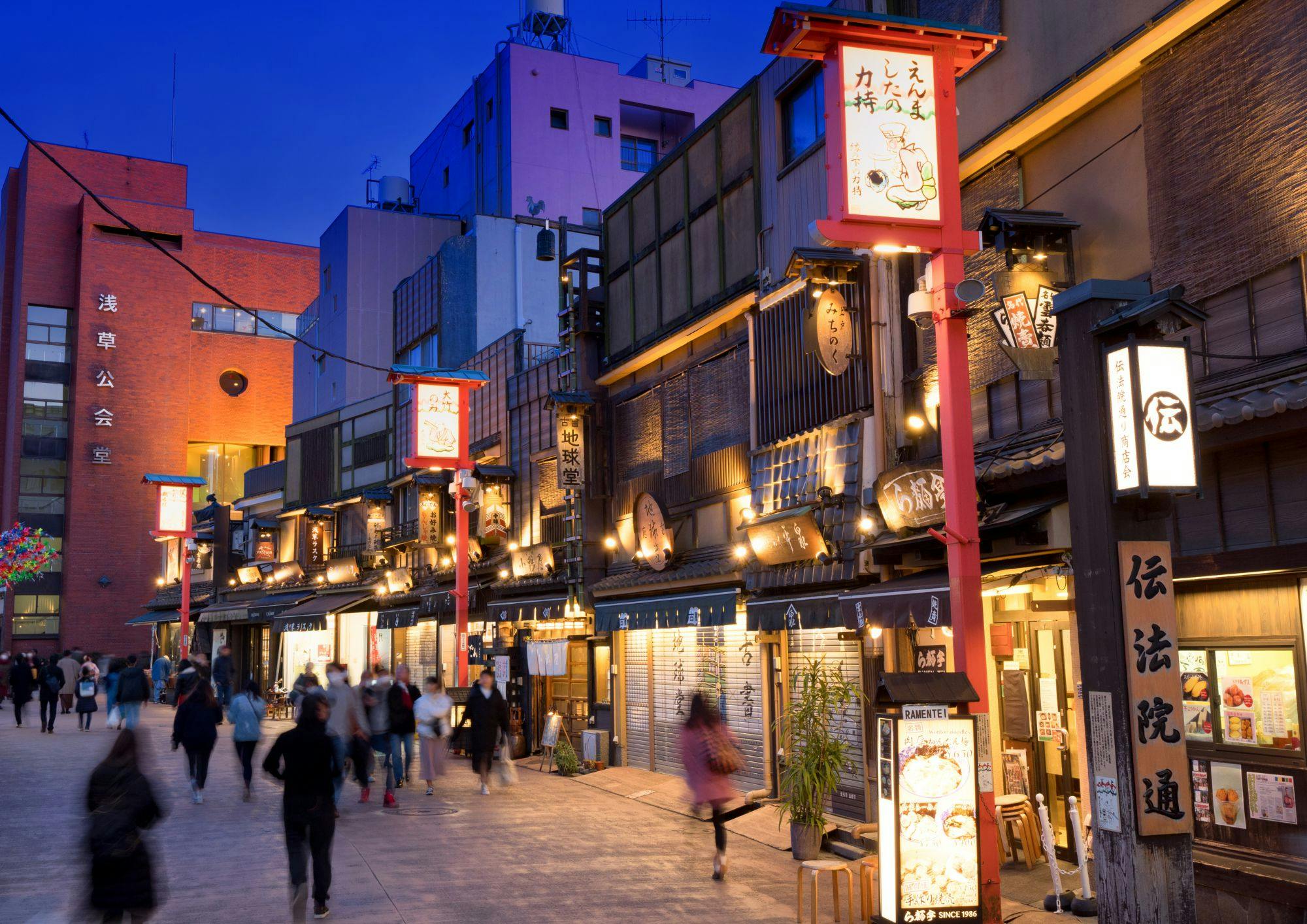 A bustling street at dusk with traditional-style shopfronts illuminated by warm lights under a deep blue sky.
