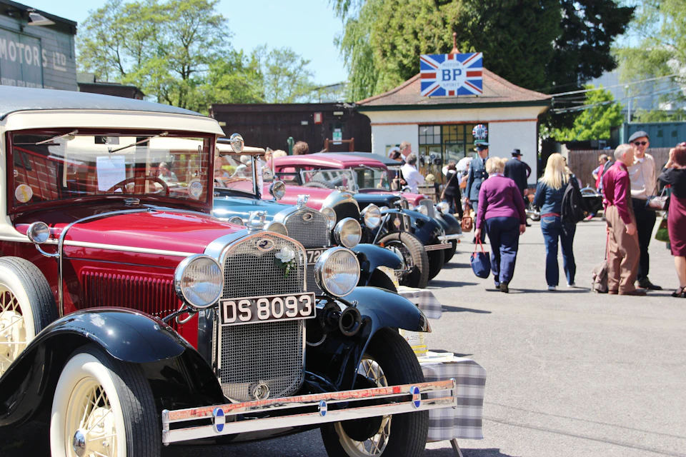 A row of vintage cars parked outside a building with a "BP" sign. People are walking and observing the cars.