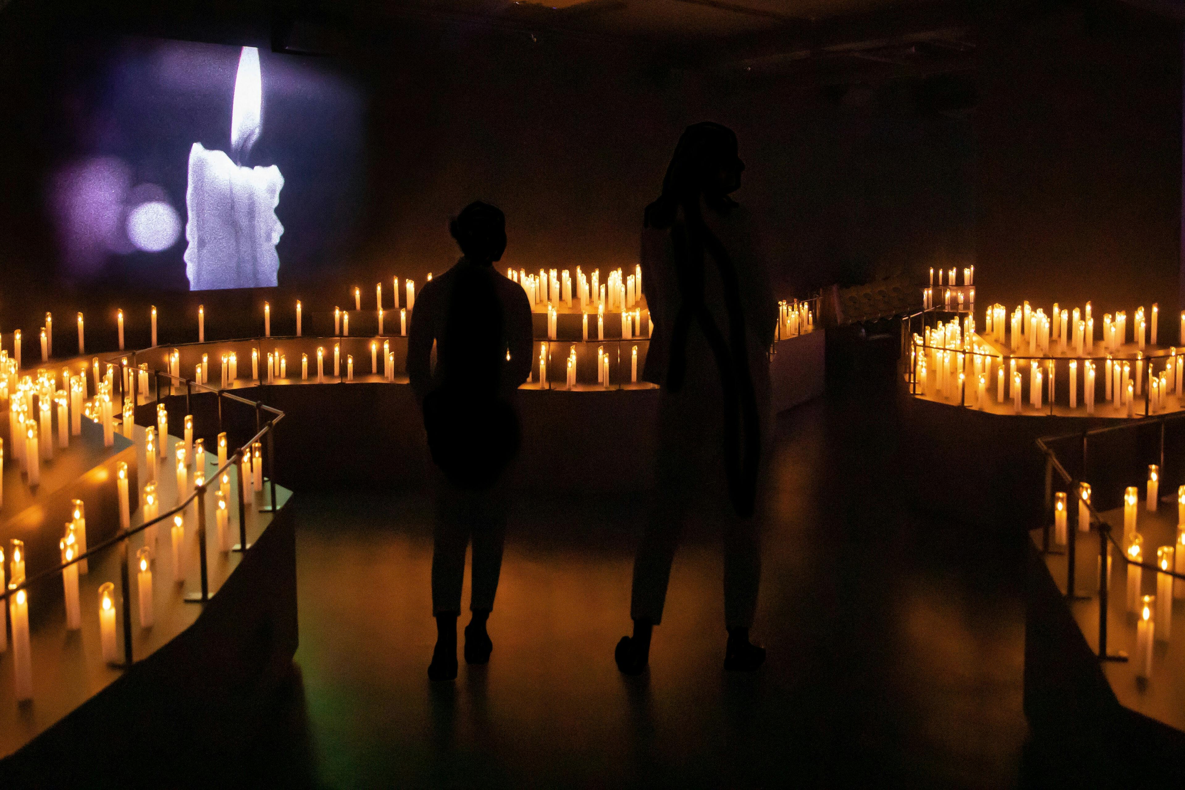 Δύο άνθρωποι στέκονται σε ένα αμυδρά φωτισμένο δωμάτιο με πολλά λαμπερά κεριά και μια προβολή ενός αναμμένου κεριού στο παρασκήνιο.