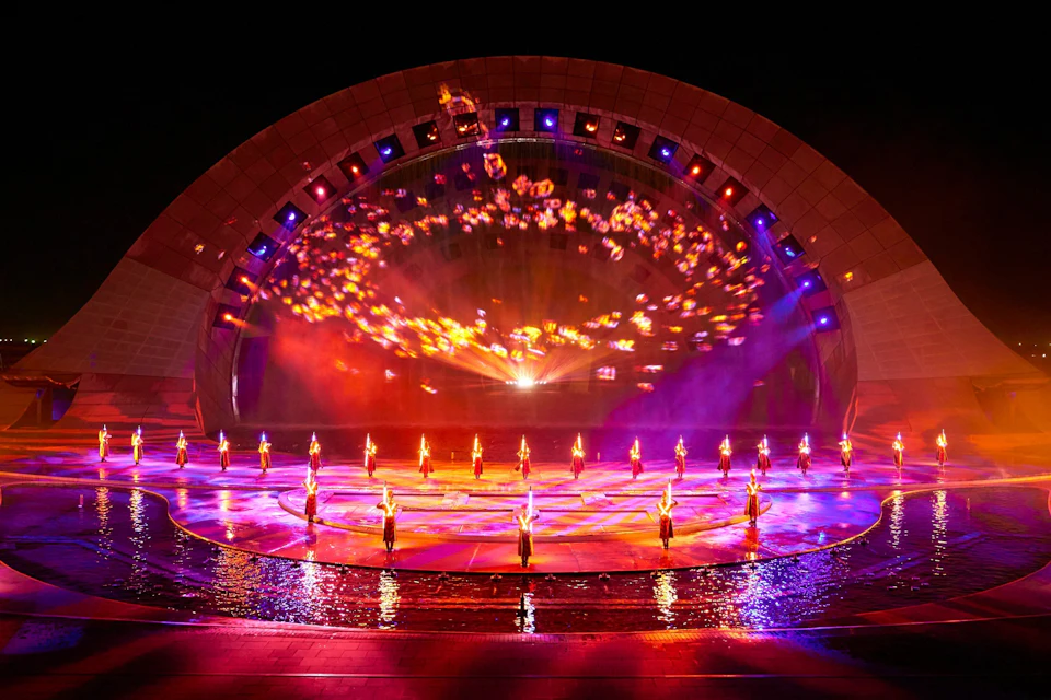 A vibrant stage performance with dancers holding lit torches, backed by a colorful light display on a large arched structure.