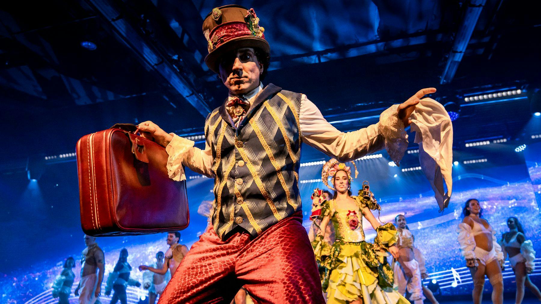 Um artista em traje vibrante com uma cartola e um saco, rodeado por outros artistas com trajes coloridos num palco iluminado.