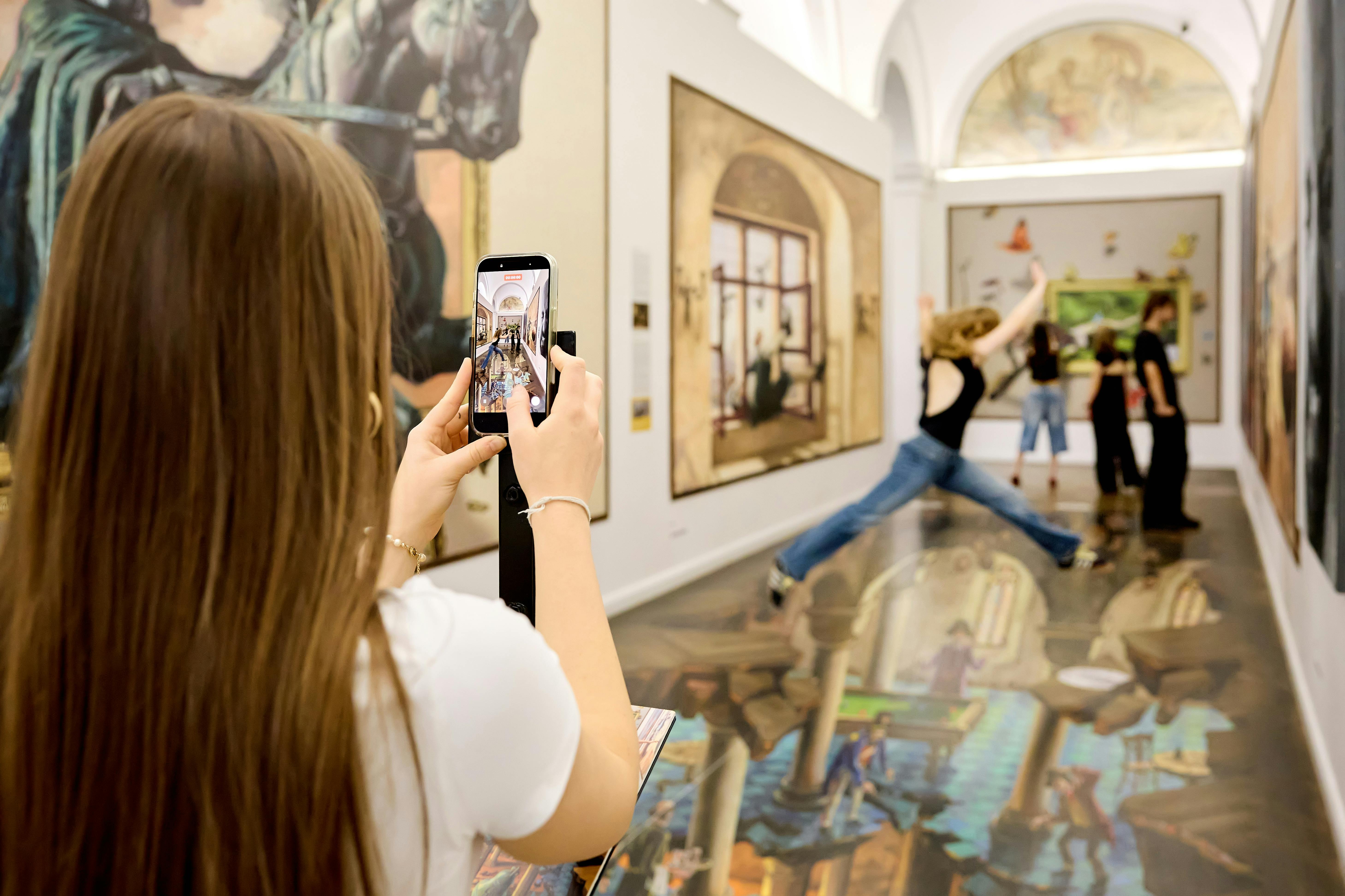 En kvinde tager et billede af et museumskunstværk med sin telefon. En person springer i baggrunden i nærheden af et stort maleri.