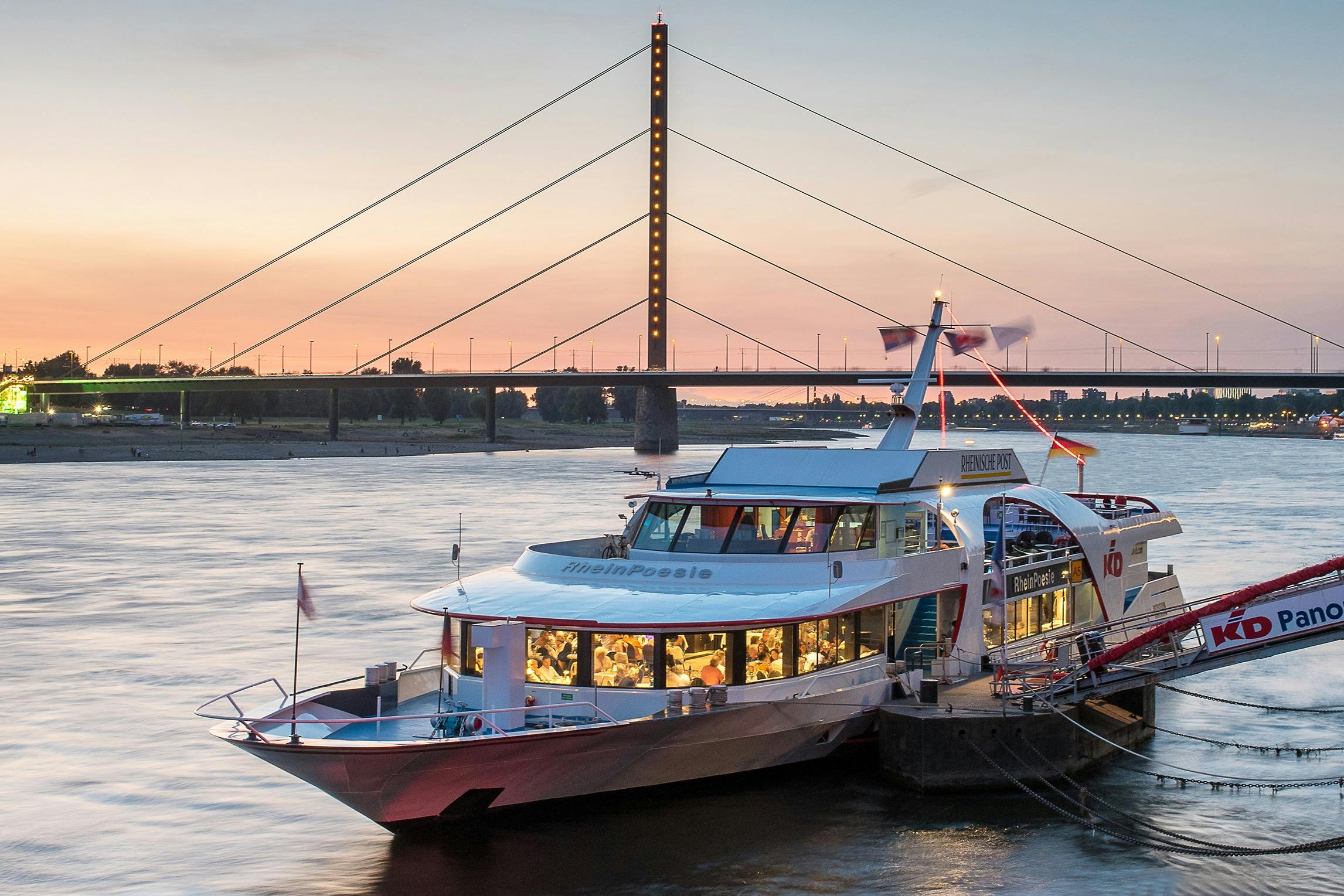 サスペンションブリッジとサンセットを背景に、ダイニングの乗客でいっぱいになったリバーボートが停泊しています。