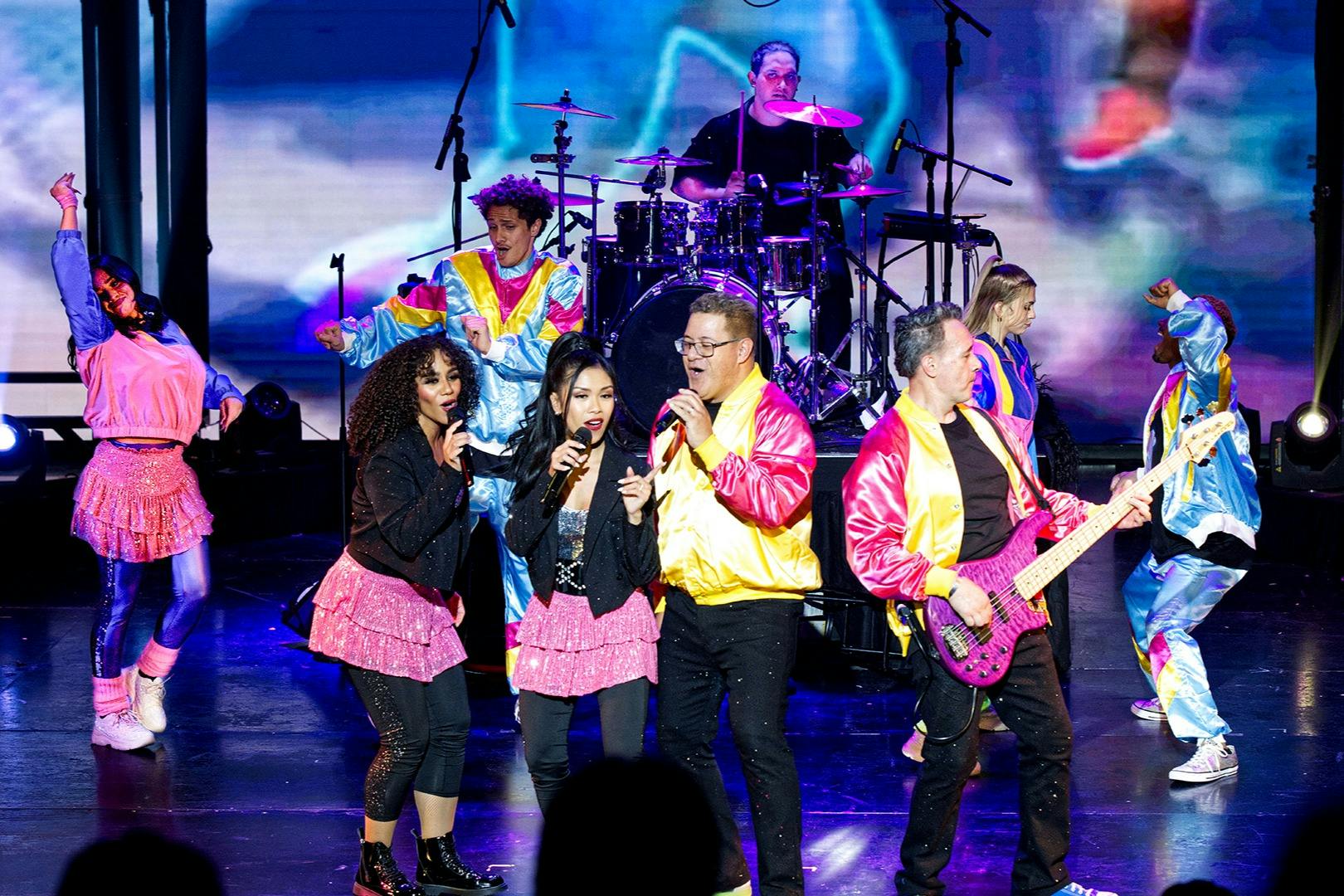 A lively stage performance featuring singers in colorful outfits, a bassist, and a drummer in the background under bright lights.