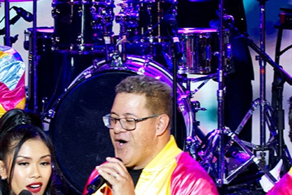 A lively stage performance featuring singers in colorful outfits, a bassist, and a drummer in the background under bright lights.